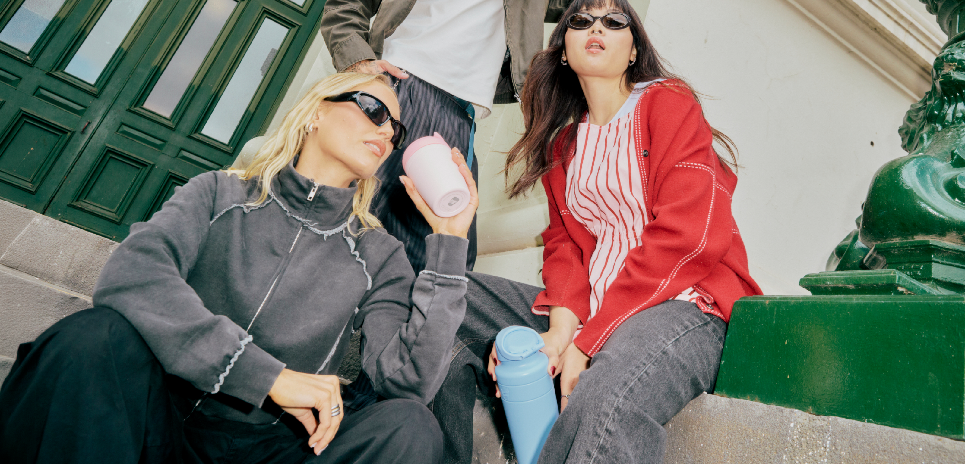 Three people sitting on outdoor stone steps wearing stylish streetwear, including sunglasses, a gray zip-up hoodie, a red cardigan over a striped top, and holding Ora insulated bottle and Commuter travel cups.