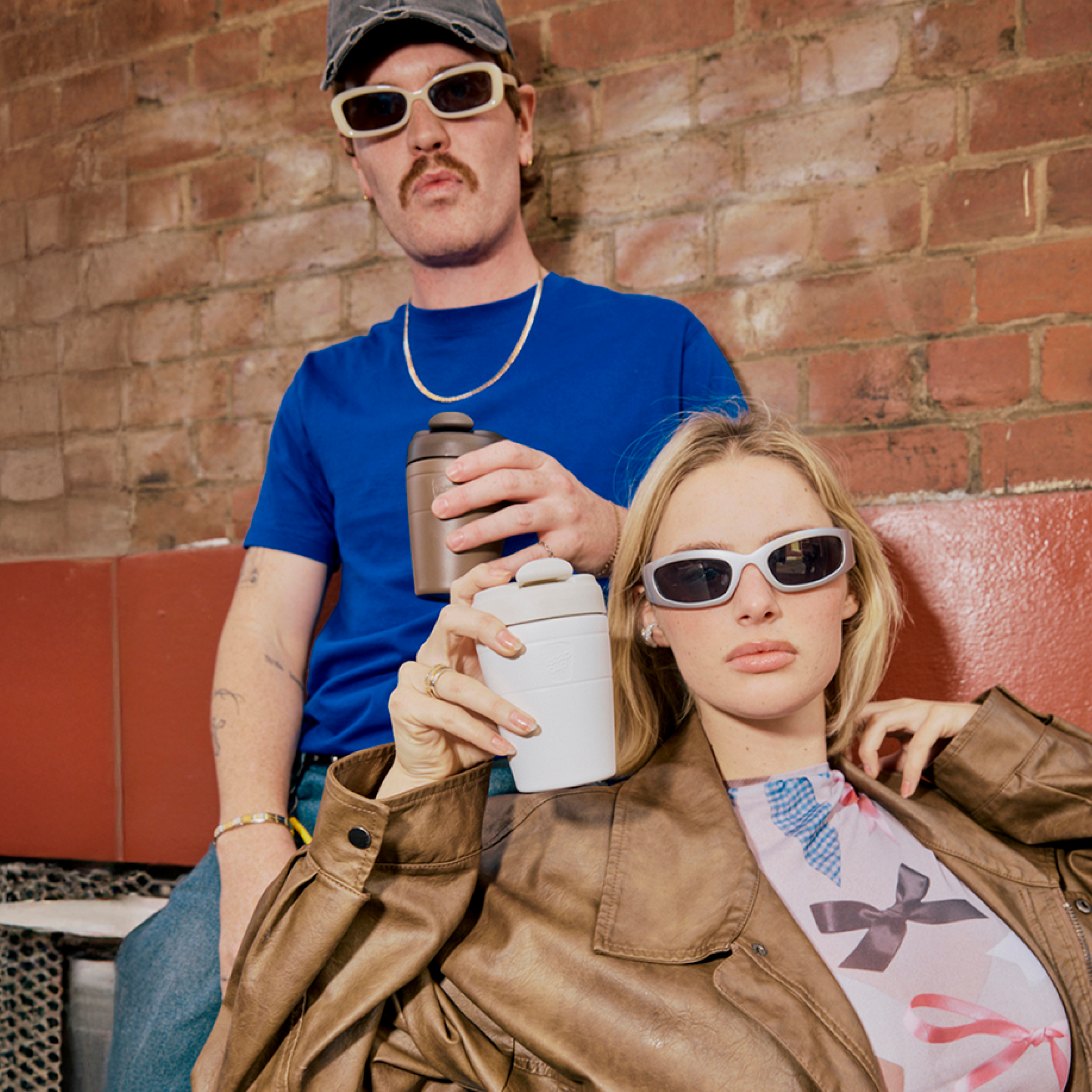 Two people wearing white sunglasses against brick wall - man in blue tee and cap, woman in brown jacket holding reusable water bottles