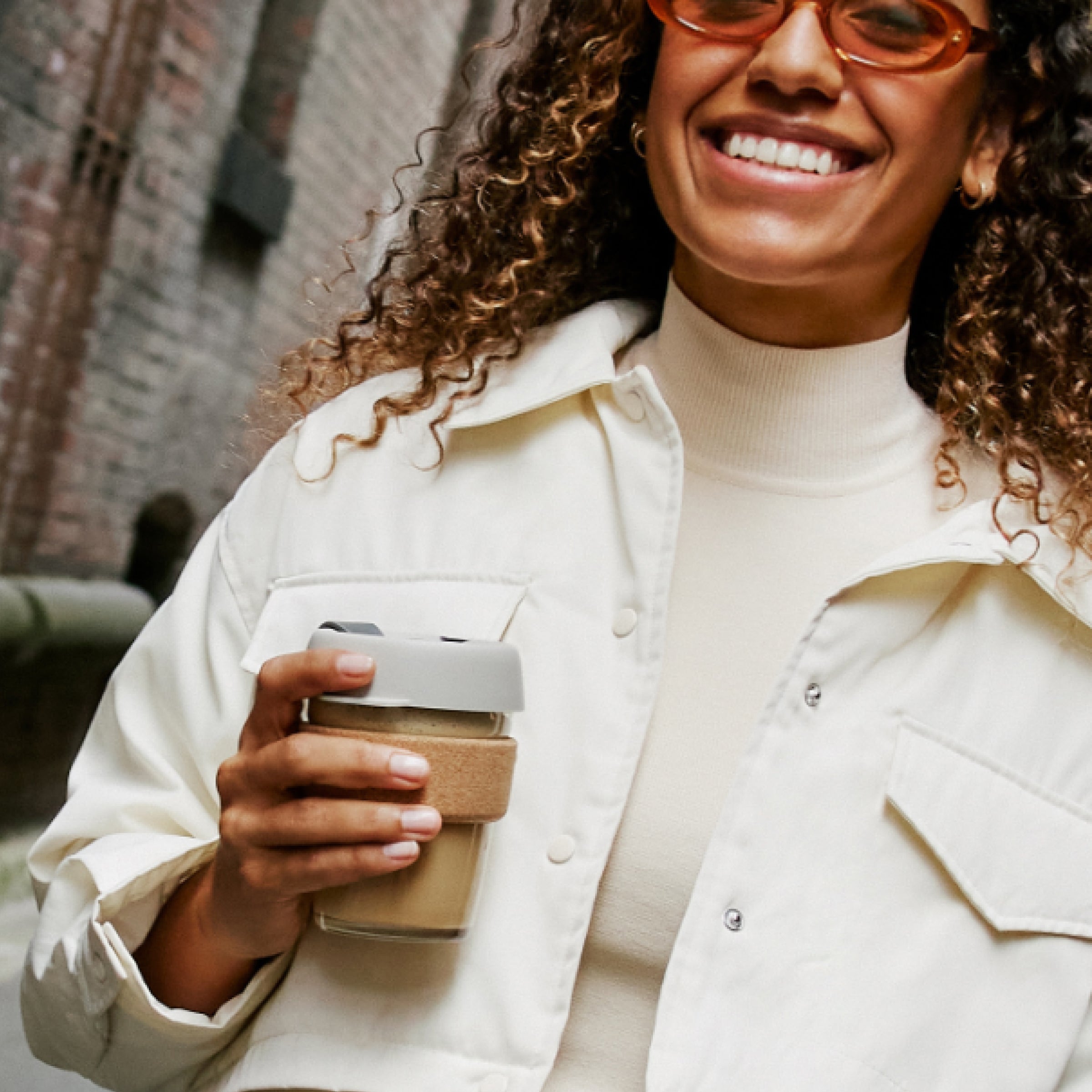 Woman with curly hair wearing cream button-up shirt and turtleneck, holding coffee cup and smiling outdoors