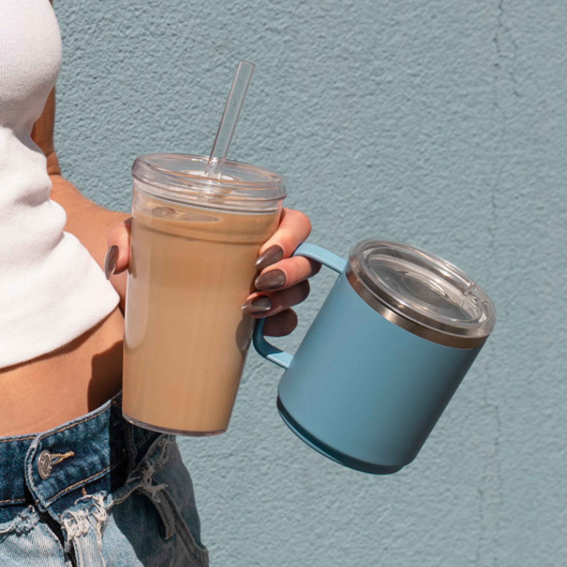 Person holding glass tumbler with iced coffee and clear straw next to blue insulated mug with clear lid against textured wall