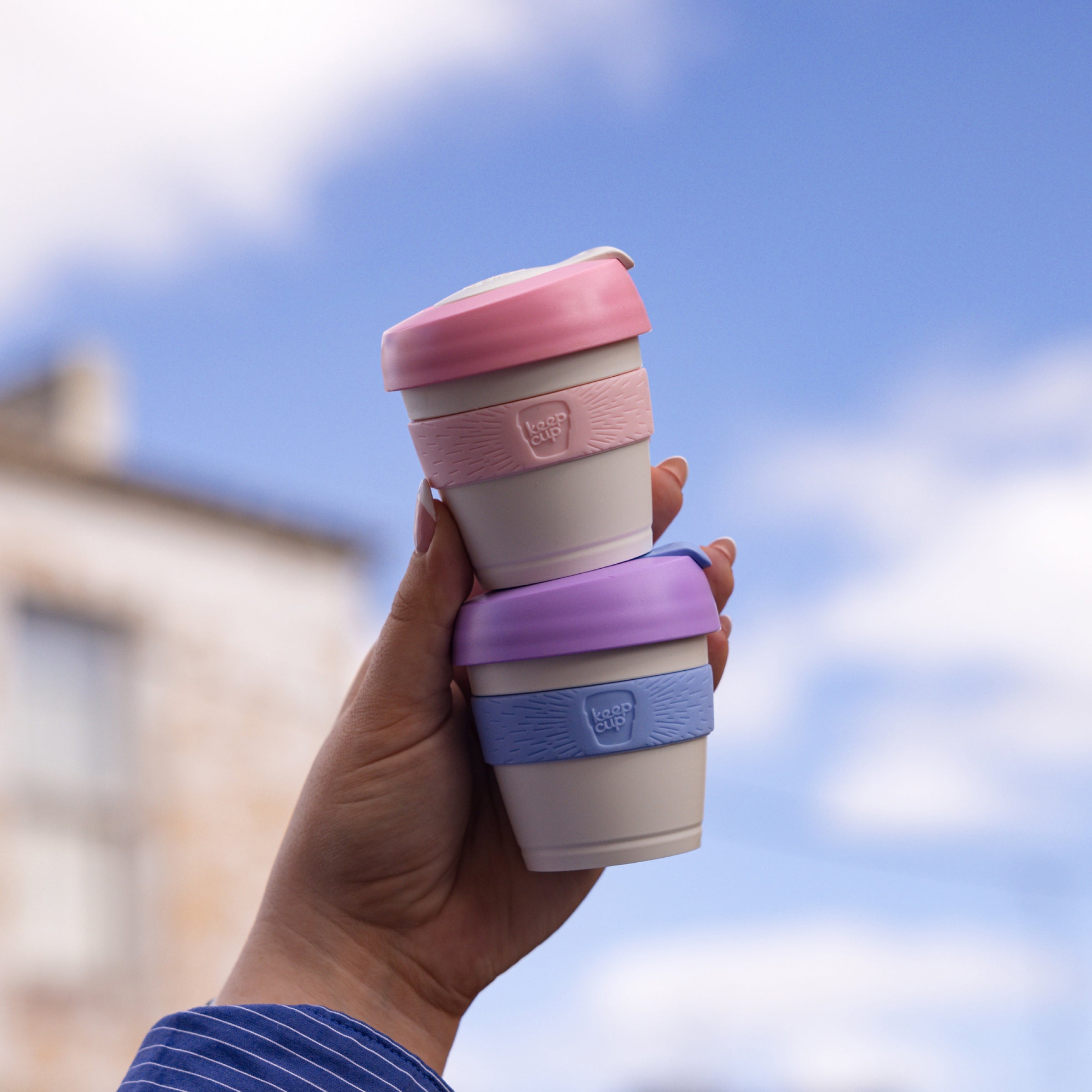 Hand holding two stacked KeepCup reusable coffee cups with pink and purple silicone bands against blue sky background