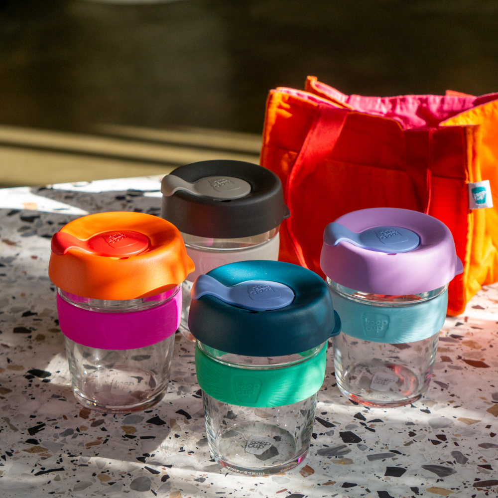 Set of glass KeepCup reusable coffee cups with colourful lids on a speckled surface with a red bag in the background.
