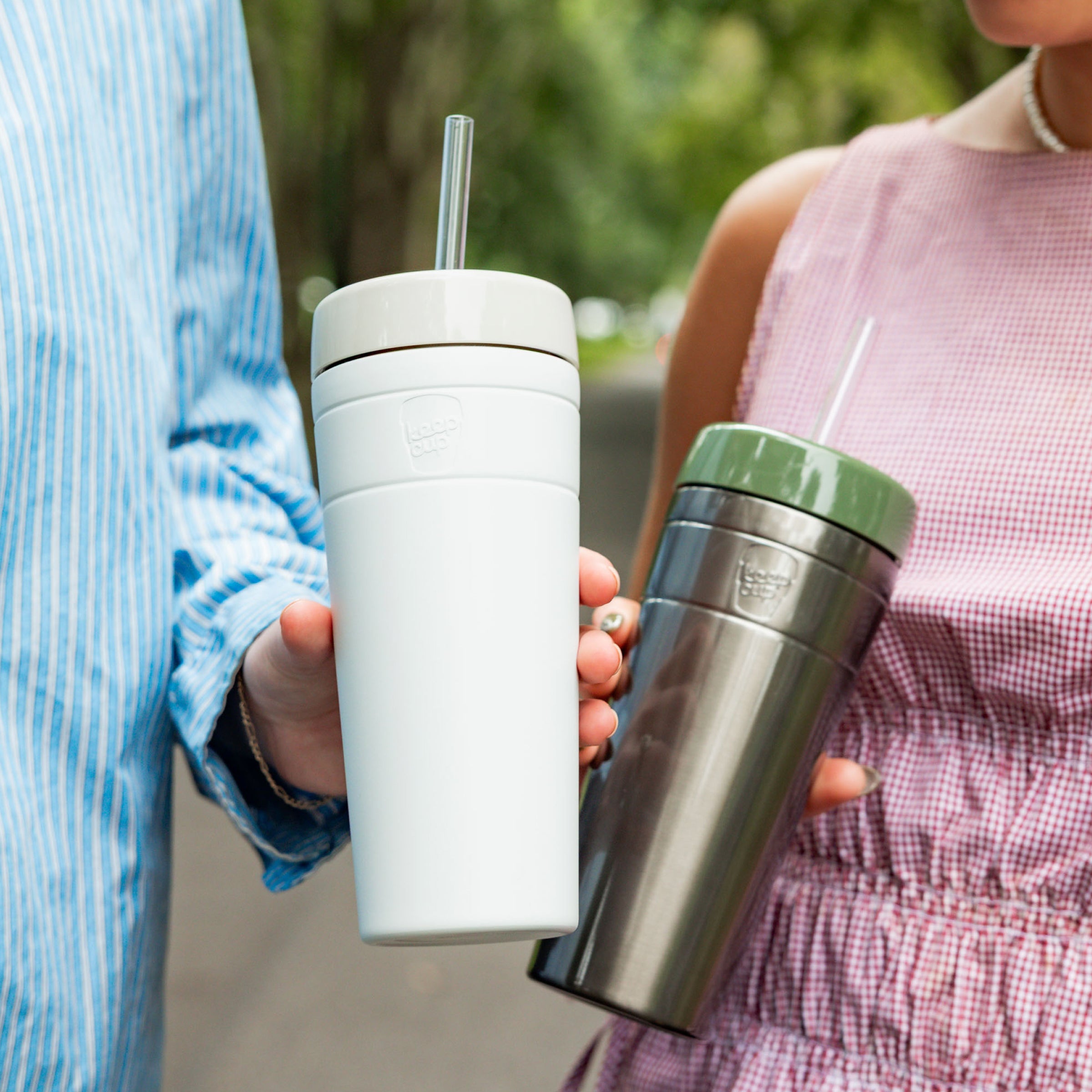 Two people outdoors holding KeepCup Commuter Cold Cups with straws — one in Bone (white with white lid) and one in stainless steel with an olive green lid — showcasing the reusable cups in a casual, social setting.