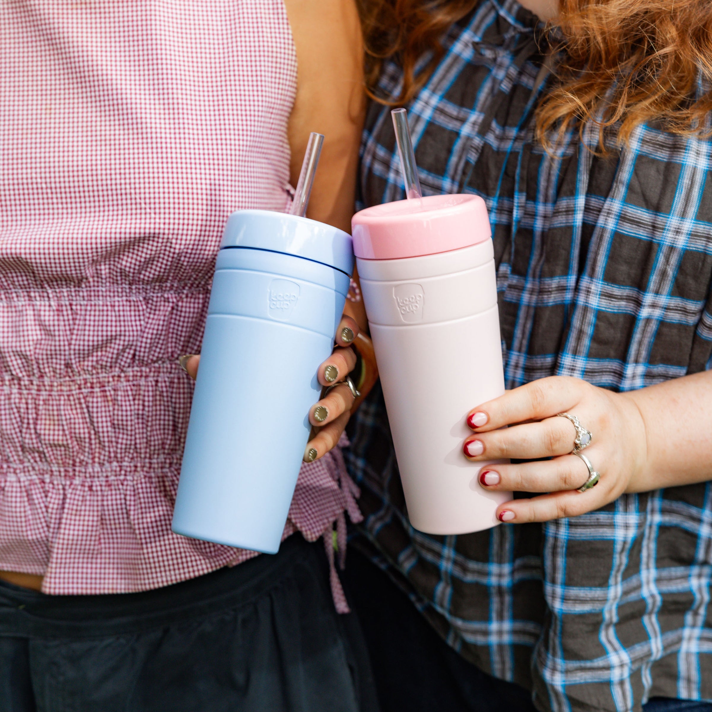 Two people standing close together, each holding a KeepCup Commuter Cold Cup with straw—one in light blue and one in Pashmak Pink—showcasing the reusable cups in a casual, friendly lifestyle setting.
