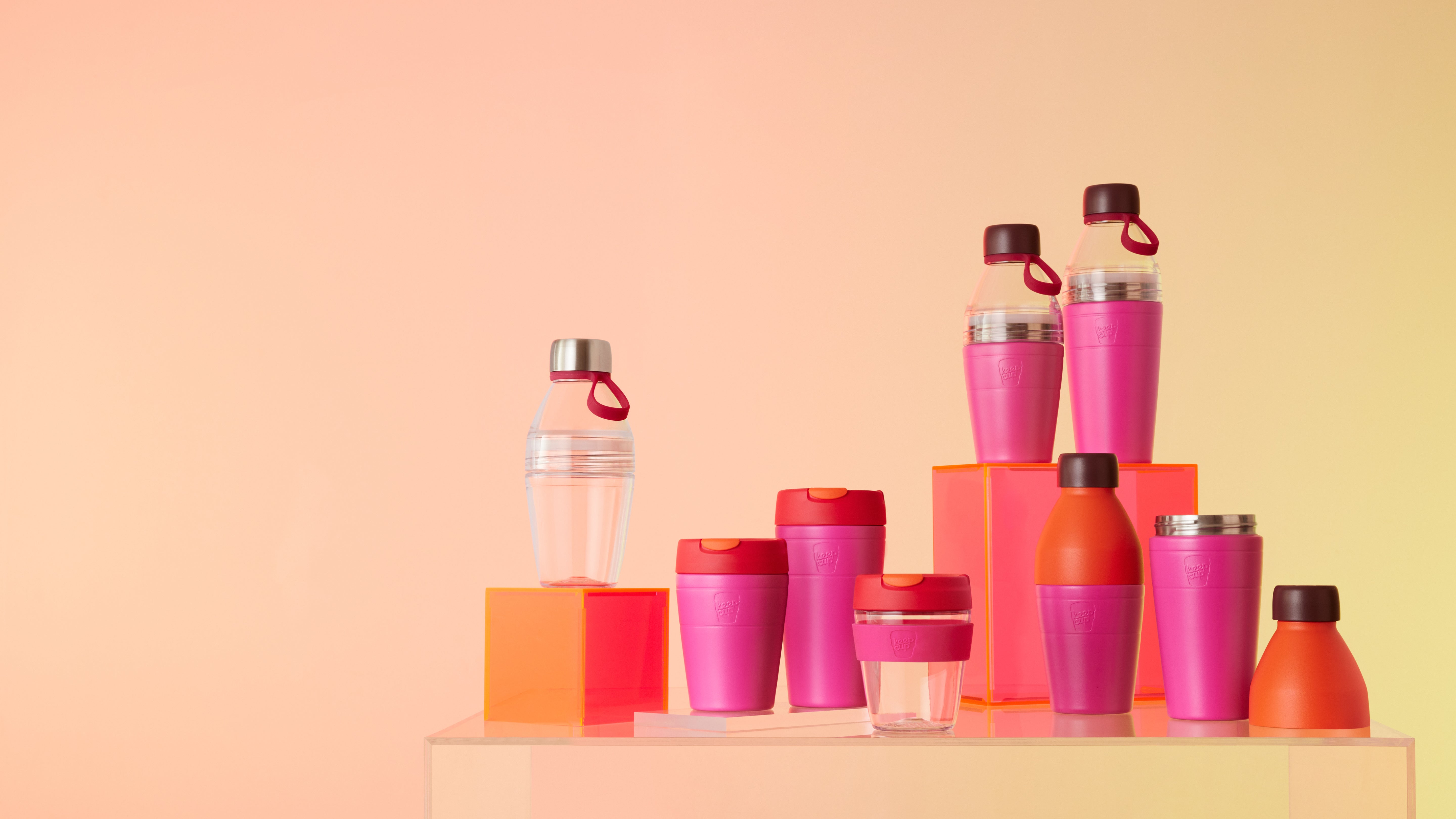 Collection of reusable drink bottles and coffee cups in pink, orange and clear colours displayed on wooden platform with geometric blocks