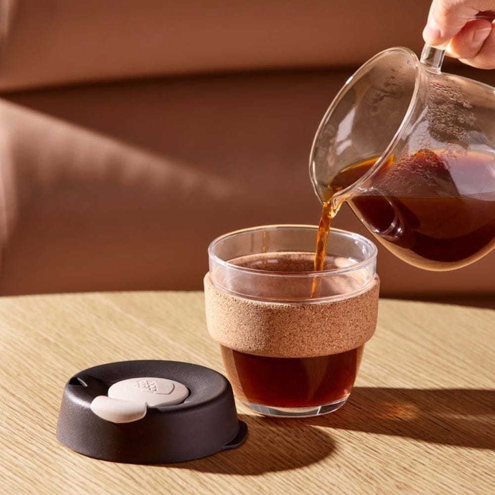 Coffee being poured from a glass server into a KeepCup Brew Cork S 8oz in Almond, showing the reusable glass cup with cork band on a wooden table, black lid with cream-coloured plug placed beside it.