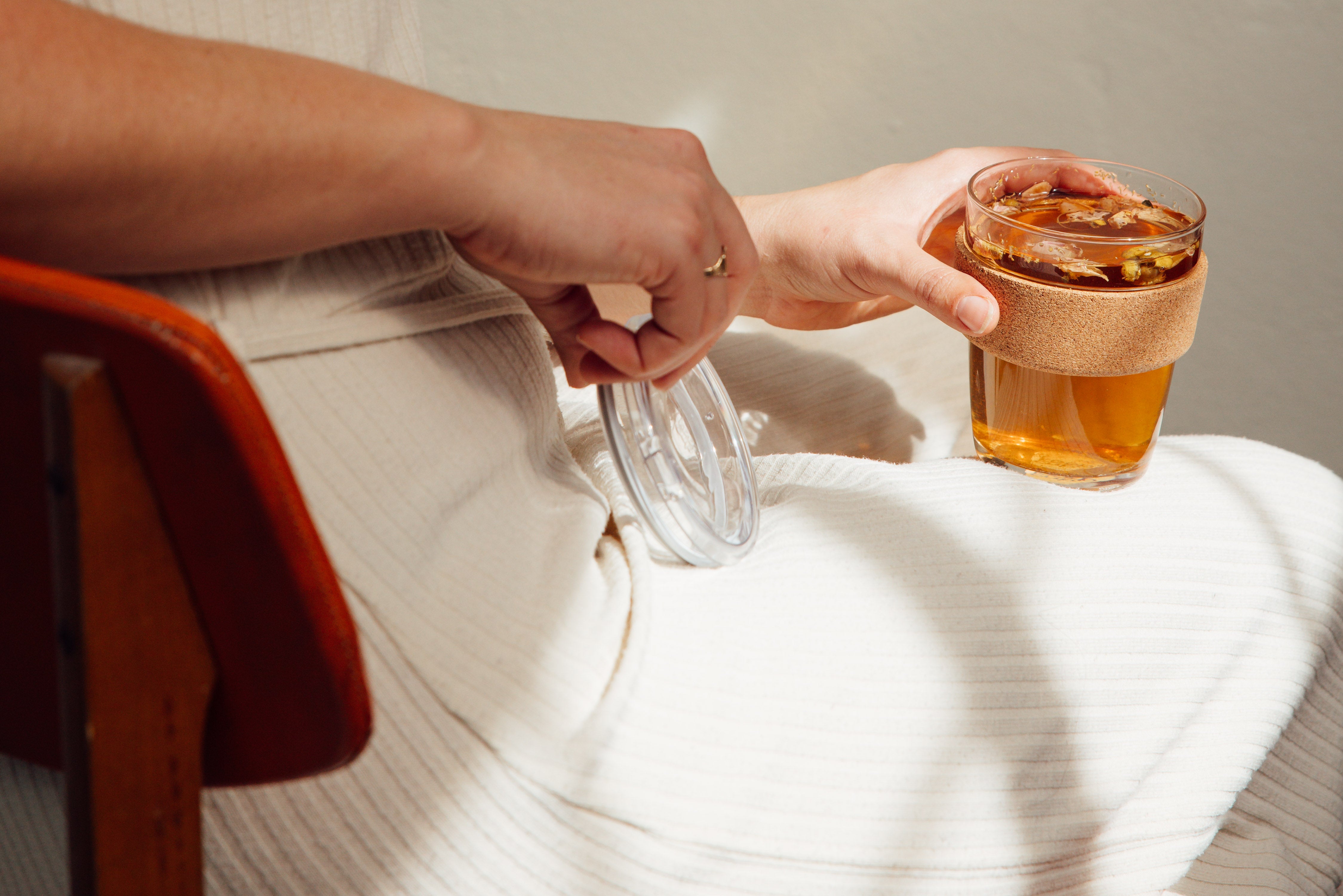 Person holding a glass of amber liquid with a slice of lemon, sitting on a chair.