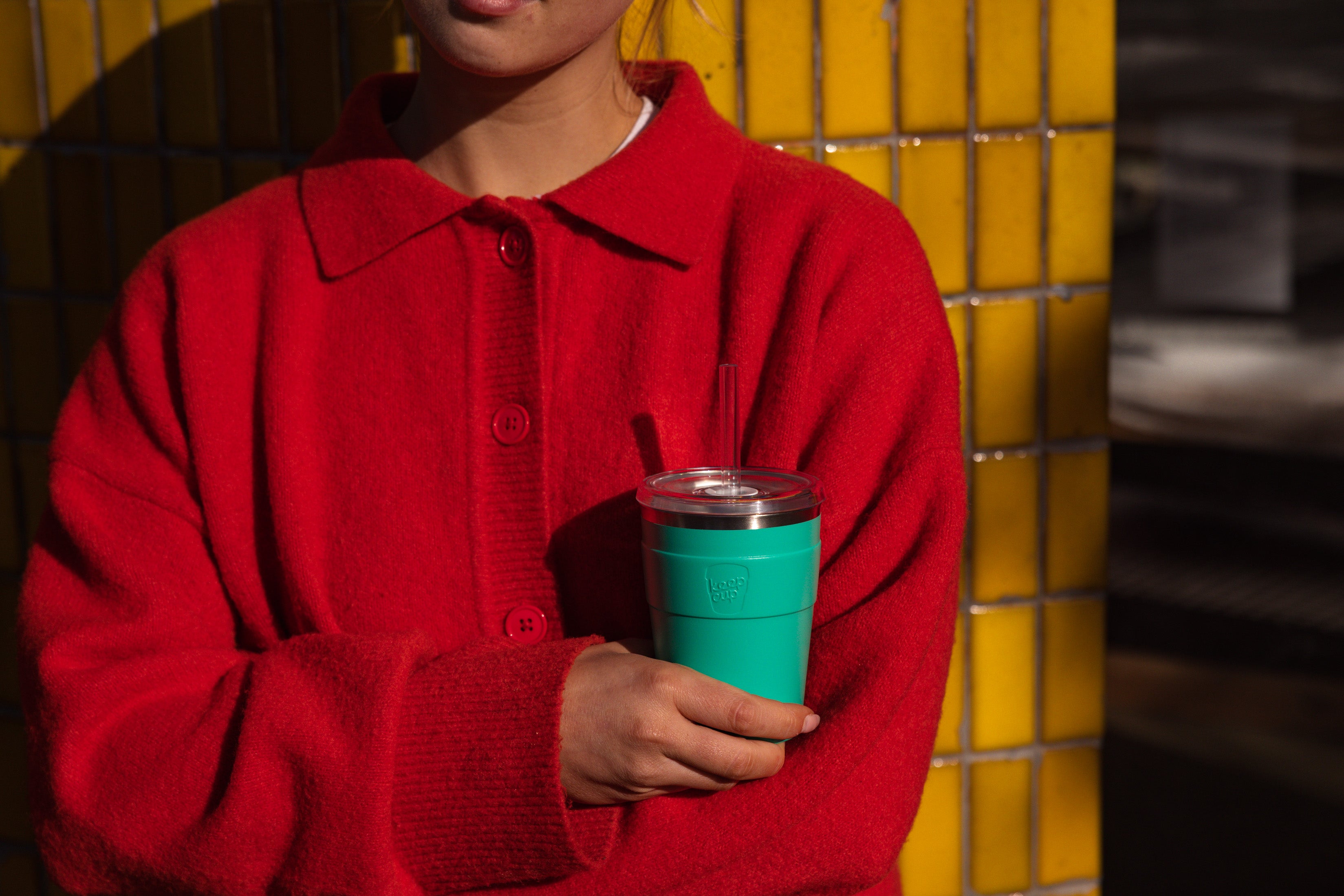 Person wearing a red sweater holding a green cup against a yellow tiled wall.