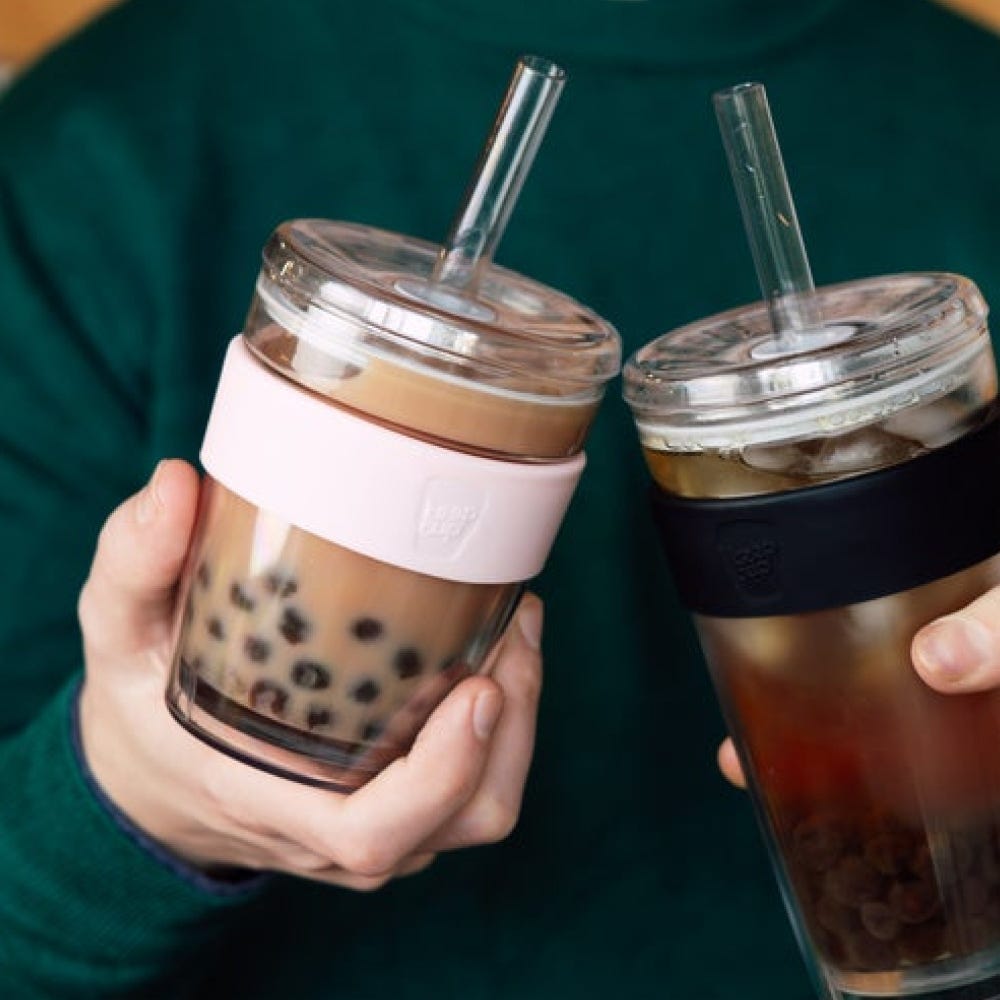 Two glass bubble tea cups with straws and silicone sleeves - one pink, one black - being held against a green background