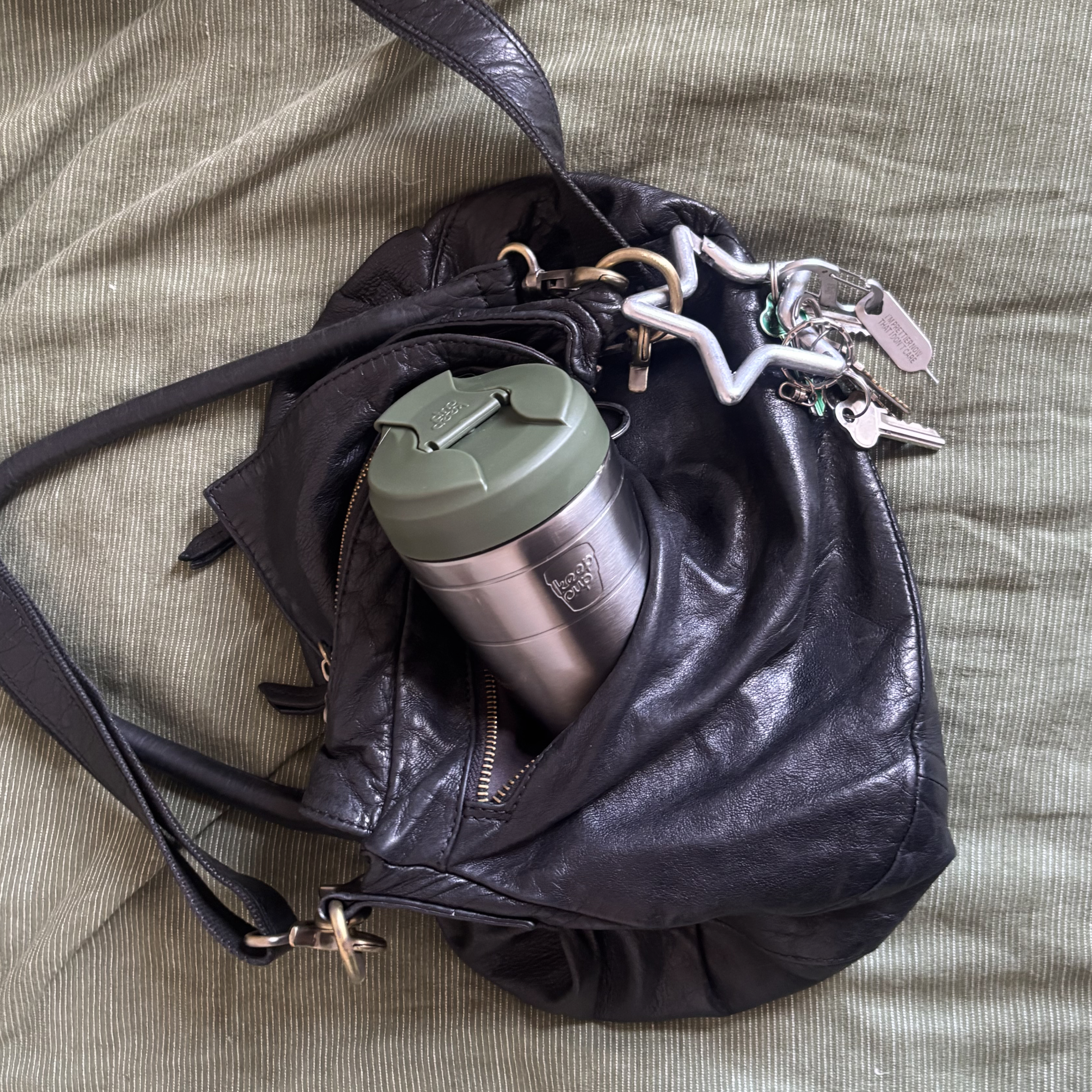 A black leather shoulder bag lying on a textured olive green fabric surface, with a silver insulated KeepCup travel mug and white keychain inserted into its side pocket.