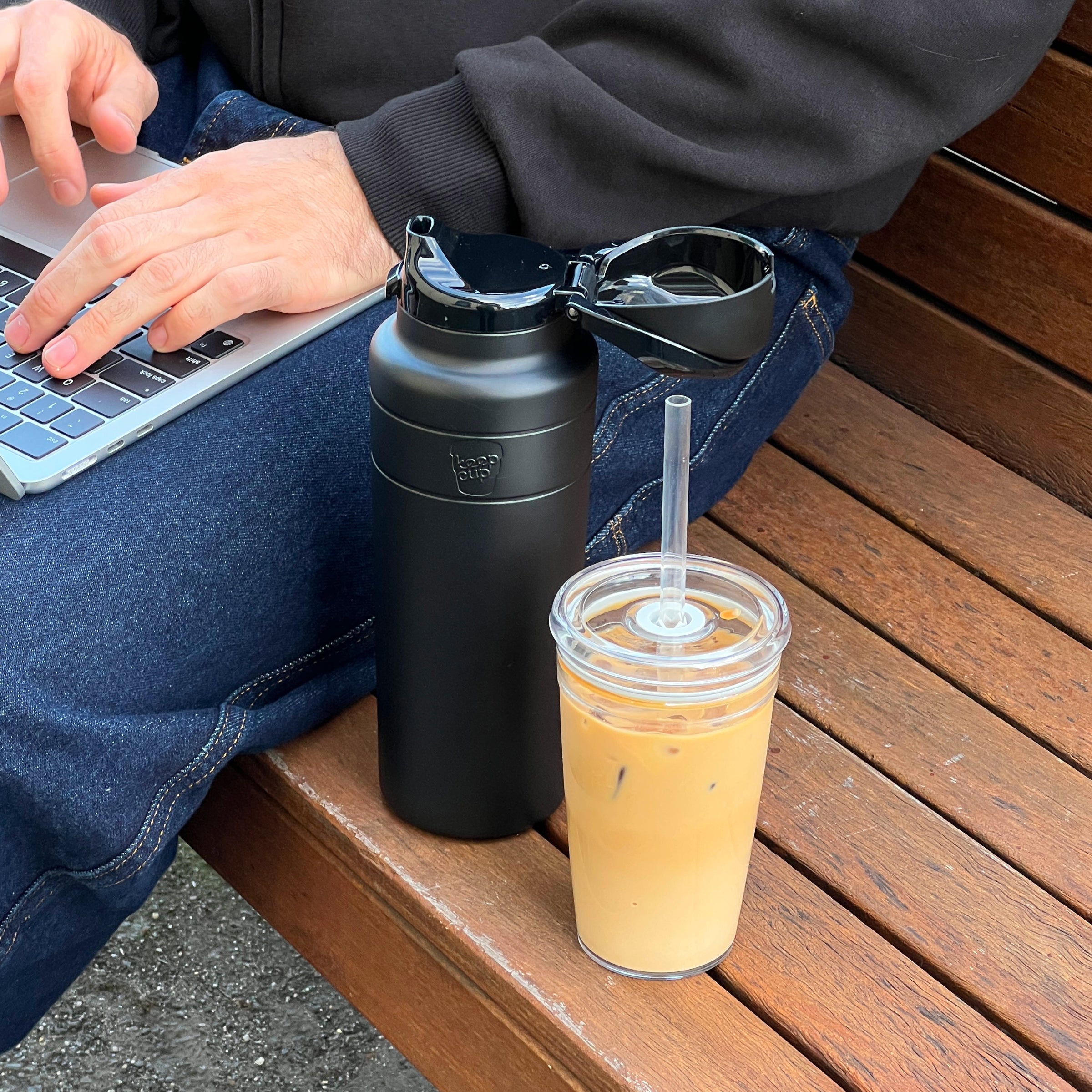 A person sits on a wooden bench working on a laptop, with a black KeepCup Ora Bottle 950ml beside them, its flip-top lid open and sunglasses resting on top, next to a clear reusable cup filled with iced coffee.