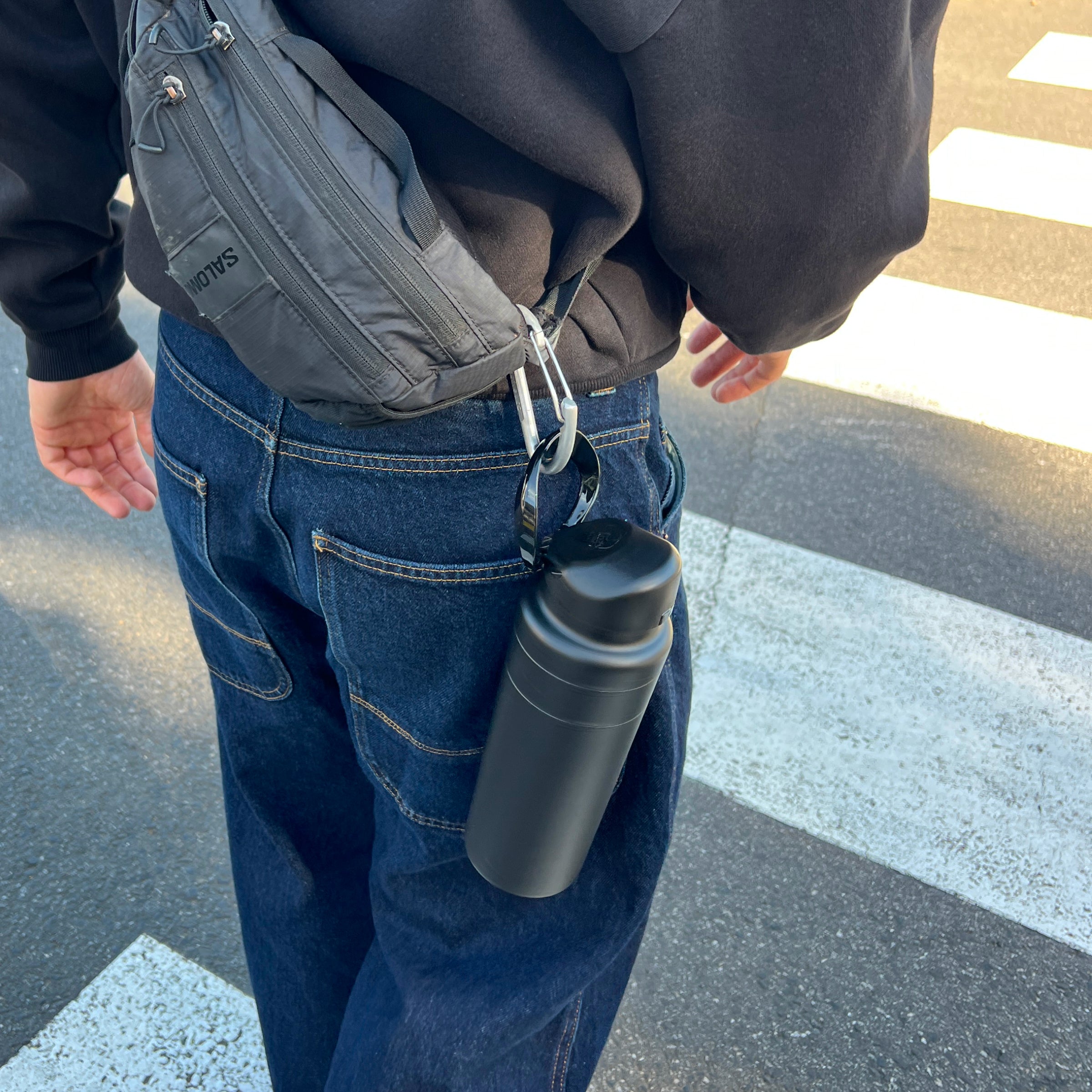 Person walking near a pedestrian crossing with a black Ora Bottle 950ml clipped to their belt loop via a carabiner, wearing dark denim jeans, a black hoodie and a grey Salomon crossbody bag.