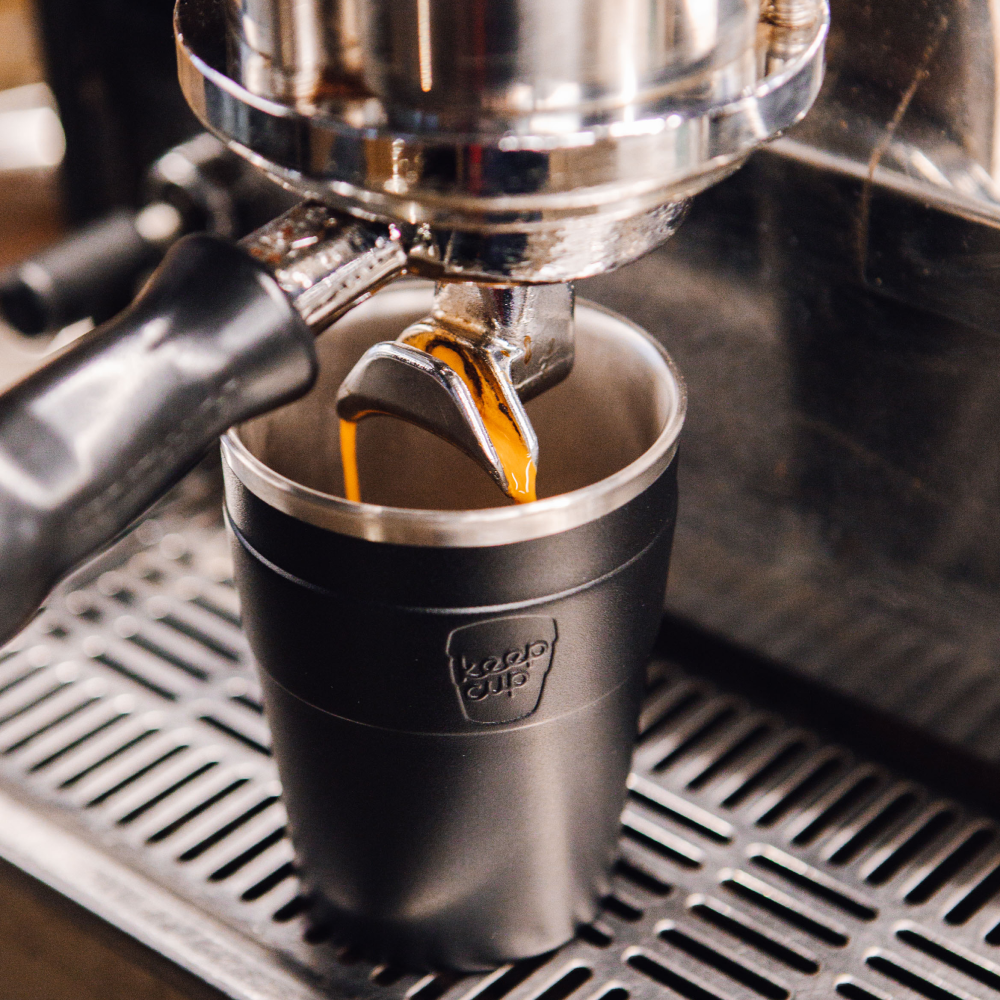 Espresso being poured into a black KeepCup stainless steel cup from a coffee machine.