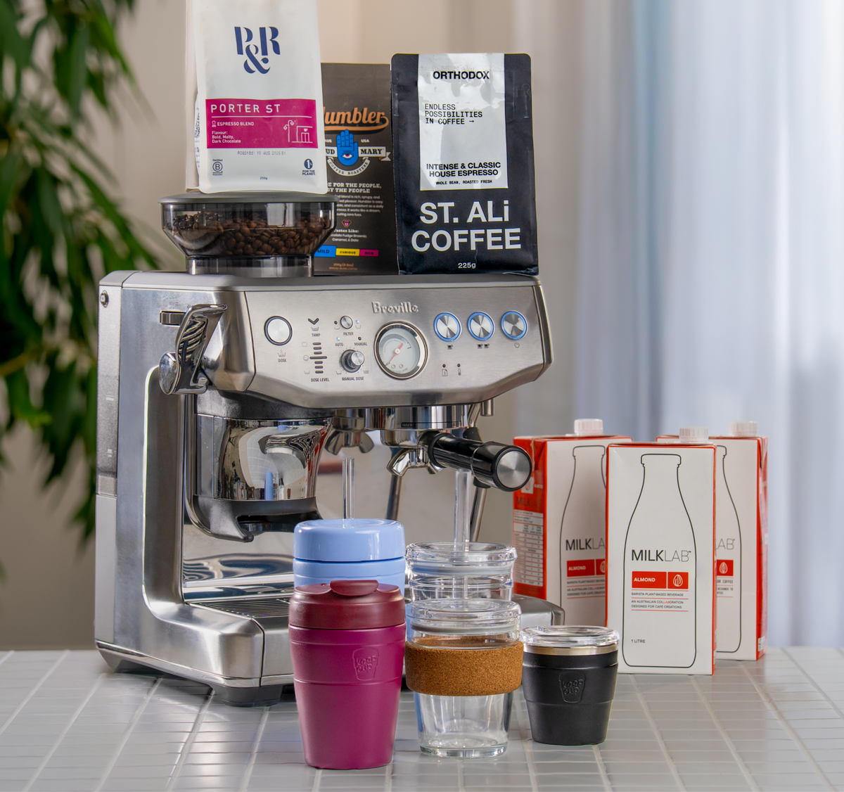 A stainless steel espresso machine on a tiled counter with coffee bags on top, milk cartons on the right, and a group of KeepCups in the front