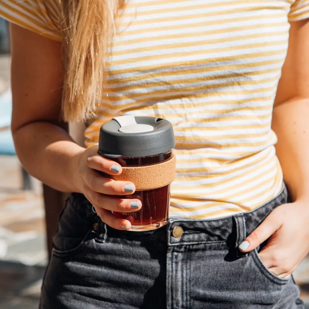 A woman in a yellow and white striped t-shirt holds a Brew Cork M 12oz KeepCup in Press colour, featuring a glass cup with cork band and dark grey lid, with black coffee visible inside.
