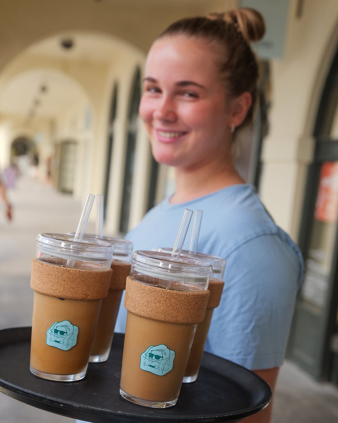 Woman holding a tray with three iced coffee drinks in clear cups with cork sleeves.