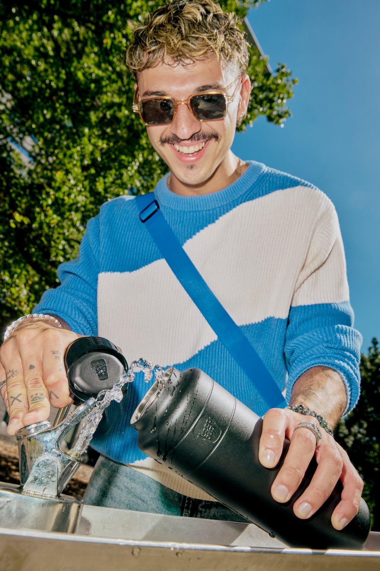 Person wearing a blue and gray sweater with sunglasses, holding a black water bottle outdoors.