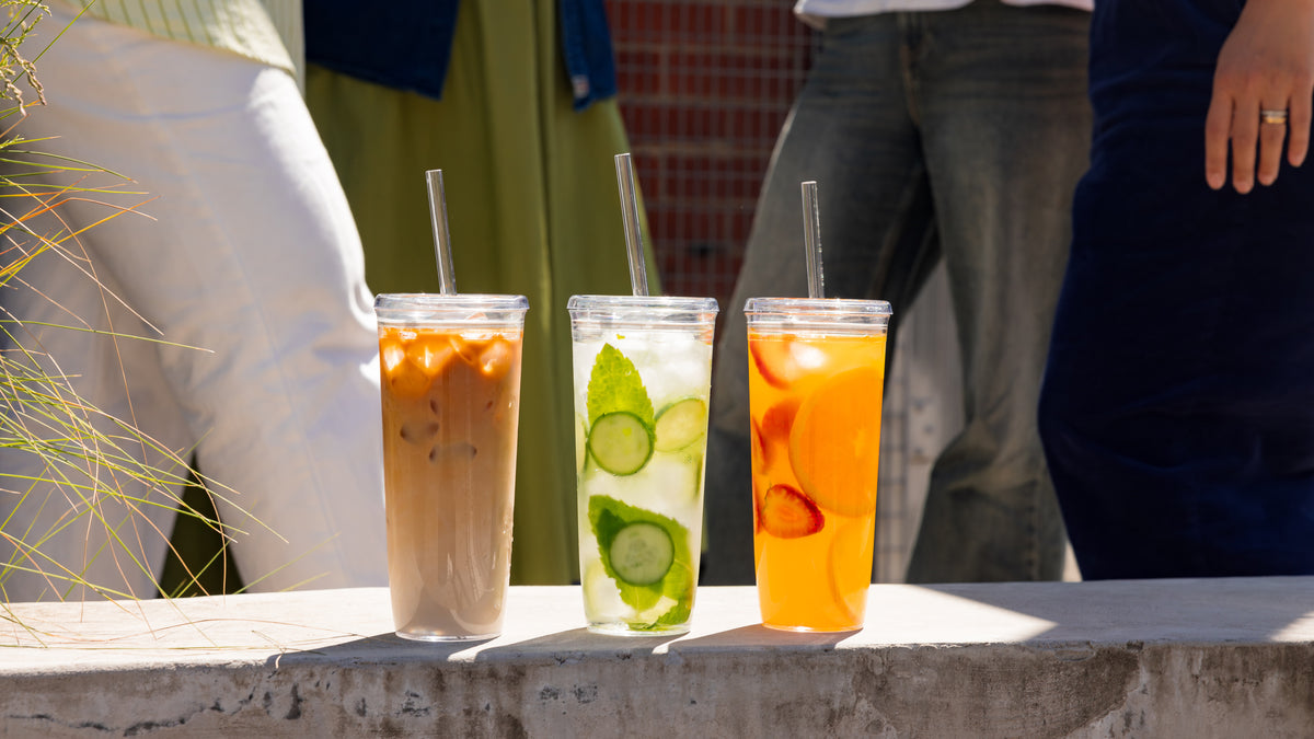 Three clear plastic cups with straws containing iced coffee, cucumber mint water, and orange fruit drink on concrete ledge