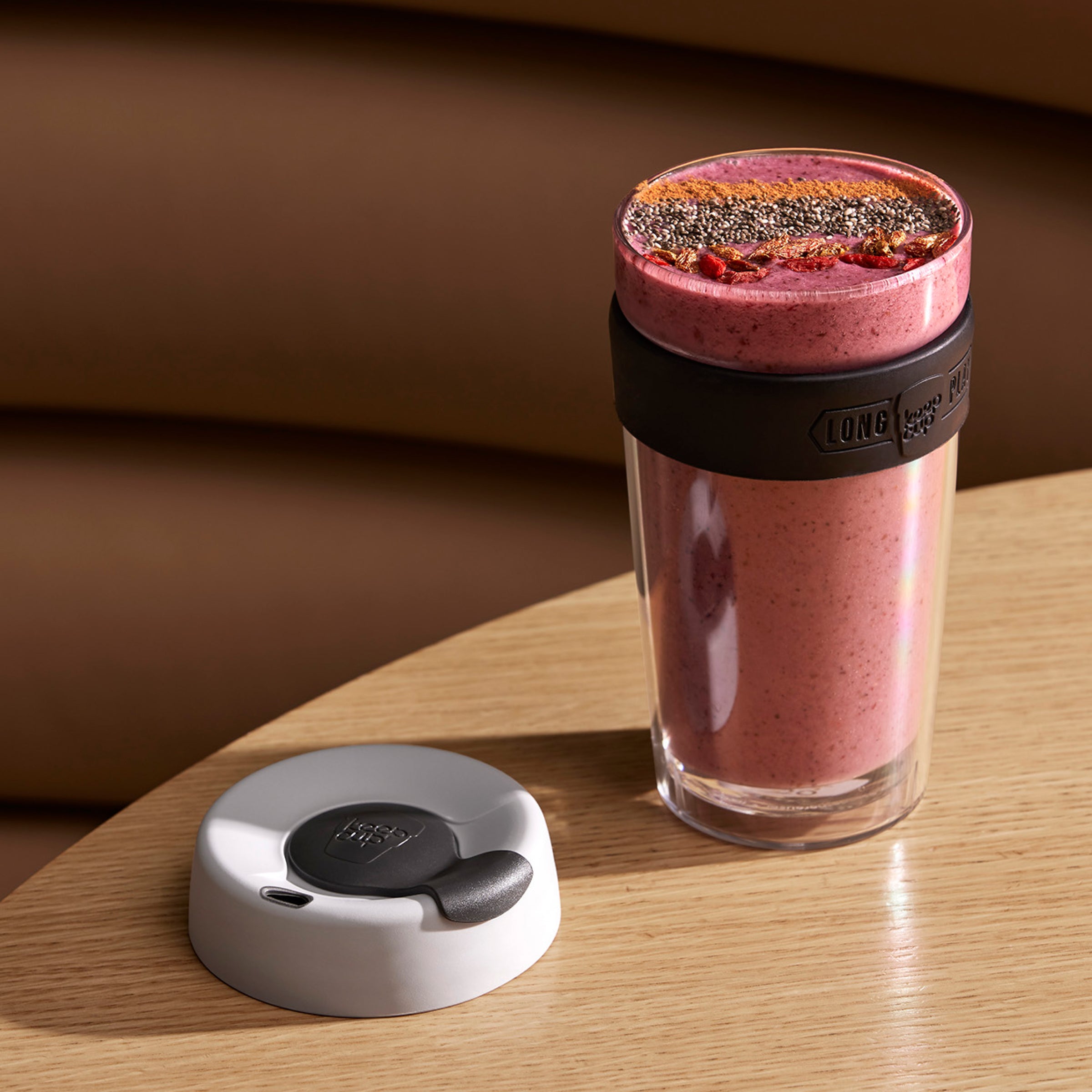 Pink smoothie in glass tumbler with black silicone band and white lid with grey spout, topped with chia seeds on wooden surface