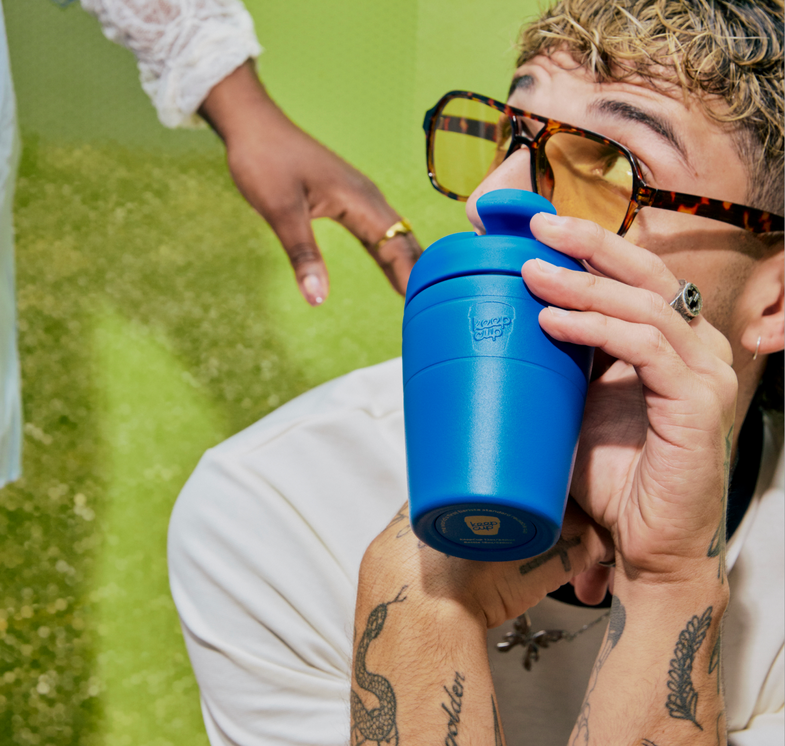 A close-up lifestyle shot of a man wearing tortoiseshell sunglasses and holding an electric blue Commuter insulated travel cup with a matching lid. 