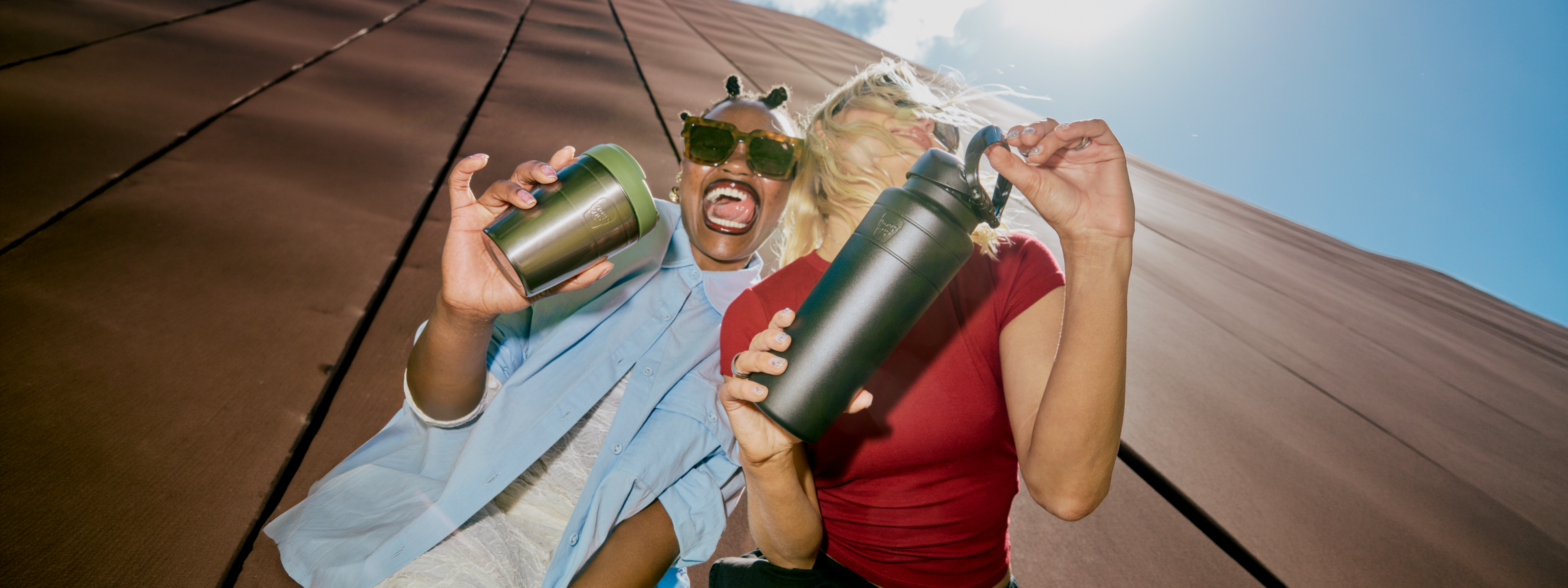 Two people outdoors on a sloped brown roof under a blue sky, one person holding a Commutre insulated travel cup and the other holdeing an Ora insulated water bottle.