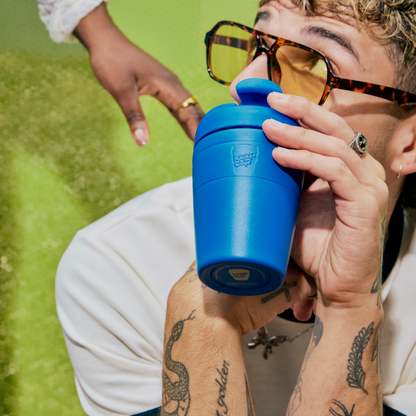 A close-up lifestyle shot of a person drinking from a bright blue reusable KeepCup Travel mug with a matching lid. The person is wearing tortoiseshell sunglasses, a ring, and has visible tattoos on their arms, set against a bright green background.