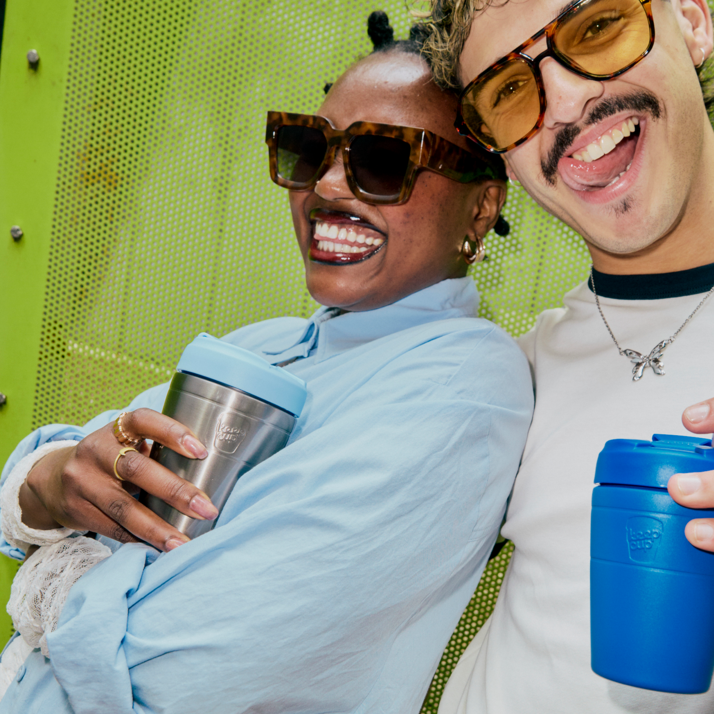 Two smiling people posing closely together in front of a bright green perforated wall, each holding a reusable KeepCup travel tumbler. The person on the left wears a light blue shirt and holds a silver tumbler with a light blue lid.