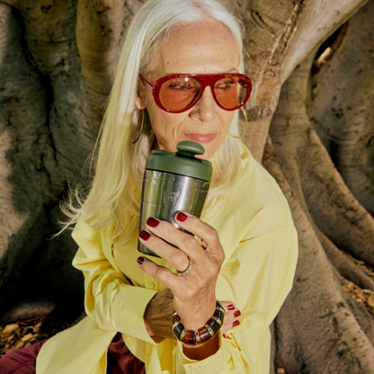 A woman with long white hair, red sunglasses, and a yellow shirt holds a stainless steel KeepCup travel tumbler with a green lid in front of a large tree trunk.