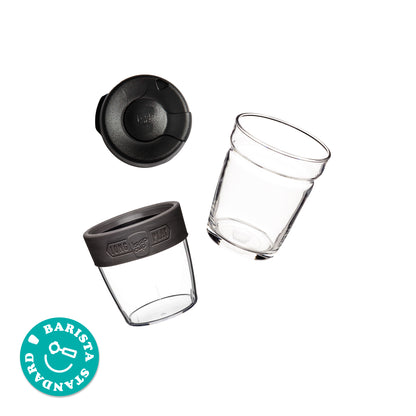 Exploded view of the KeepCup Brew Longplay M 12oz showing the splashproof lid, glass cup with charcoal-coloured band, and taller plastic outer layer separately, alongside a circular sticker reading "Barista Standard".