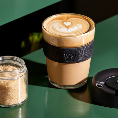 Cappuccino in a KeepCup reusable coffee cup with a black sleeve with a black lid on a green surface, next to a jar of sugar.