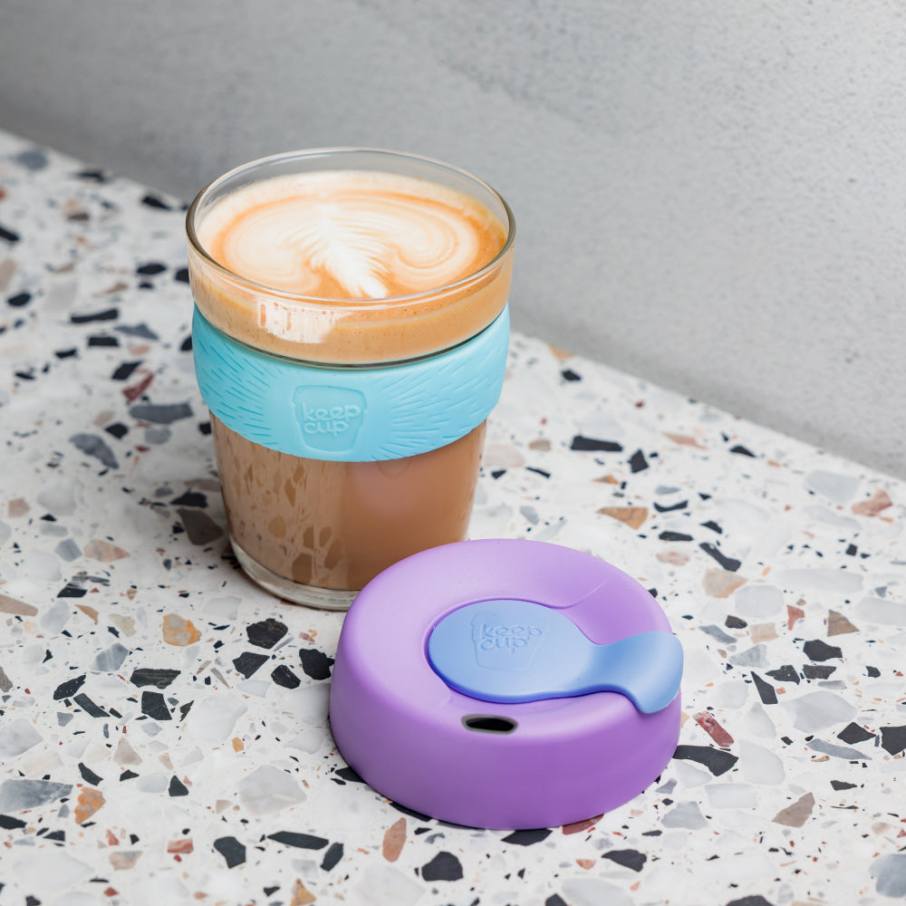 Cappuccino in a KeepCup glass reusable coffee up with a blue sleeve and purple lid on a terrazzo countertop.
