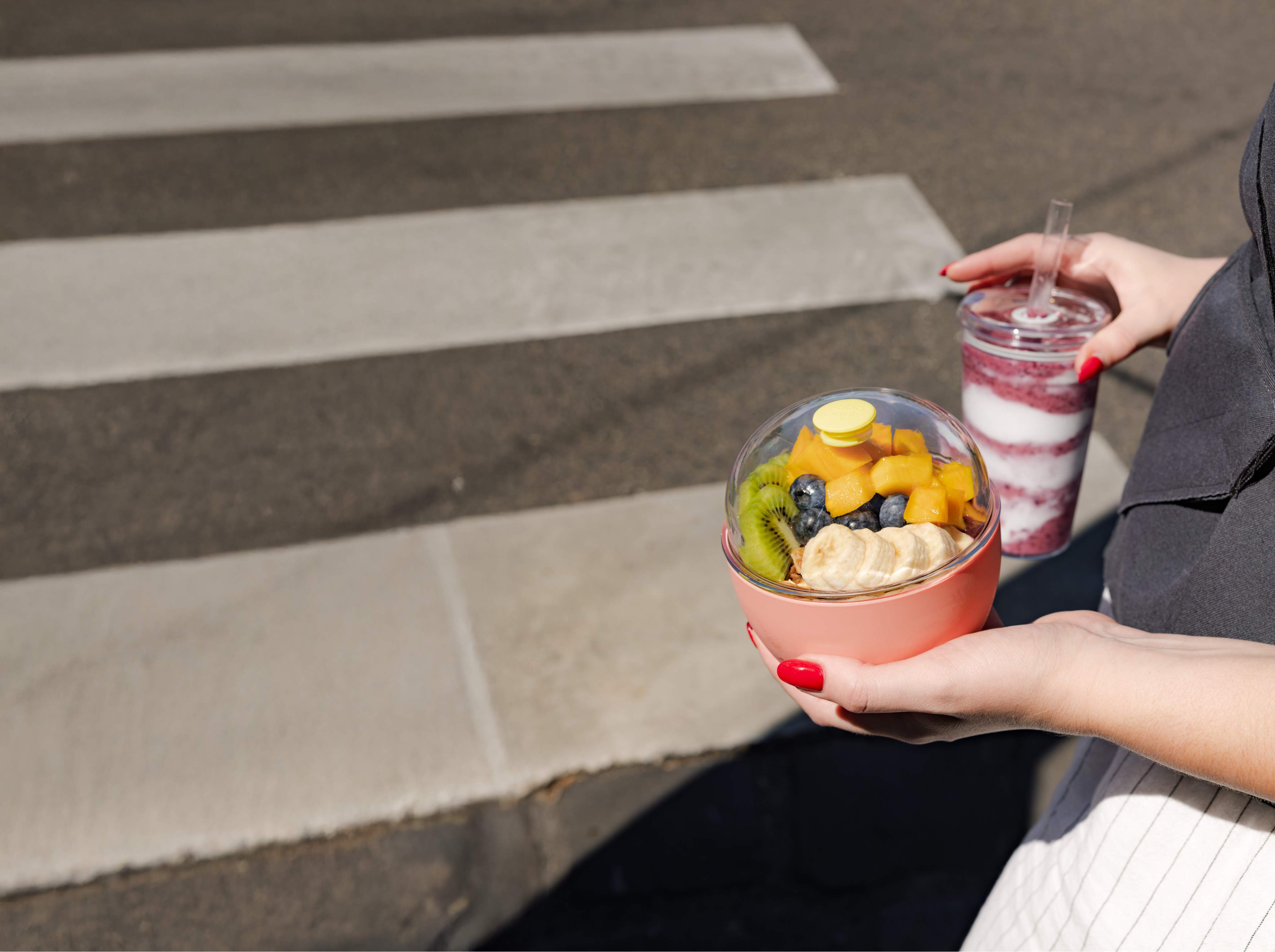 Person holding a bowl of fruit and a drink on a crosswalk