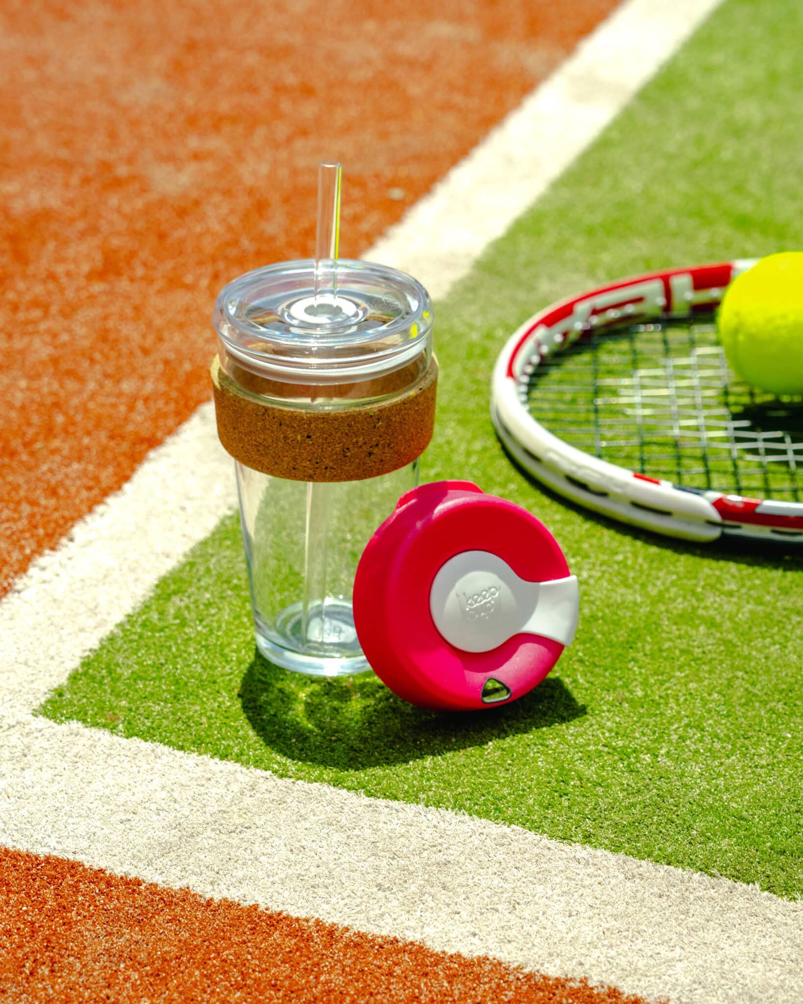 Glass water bottle with brown cork grip sleeve and red lid on tennis court beside racquet