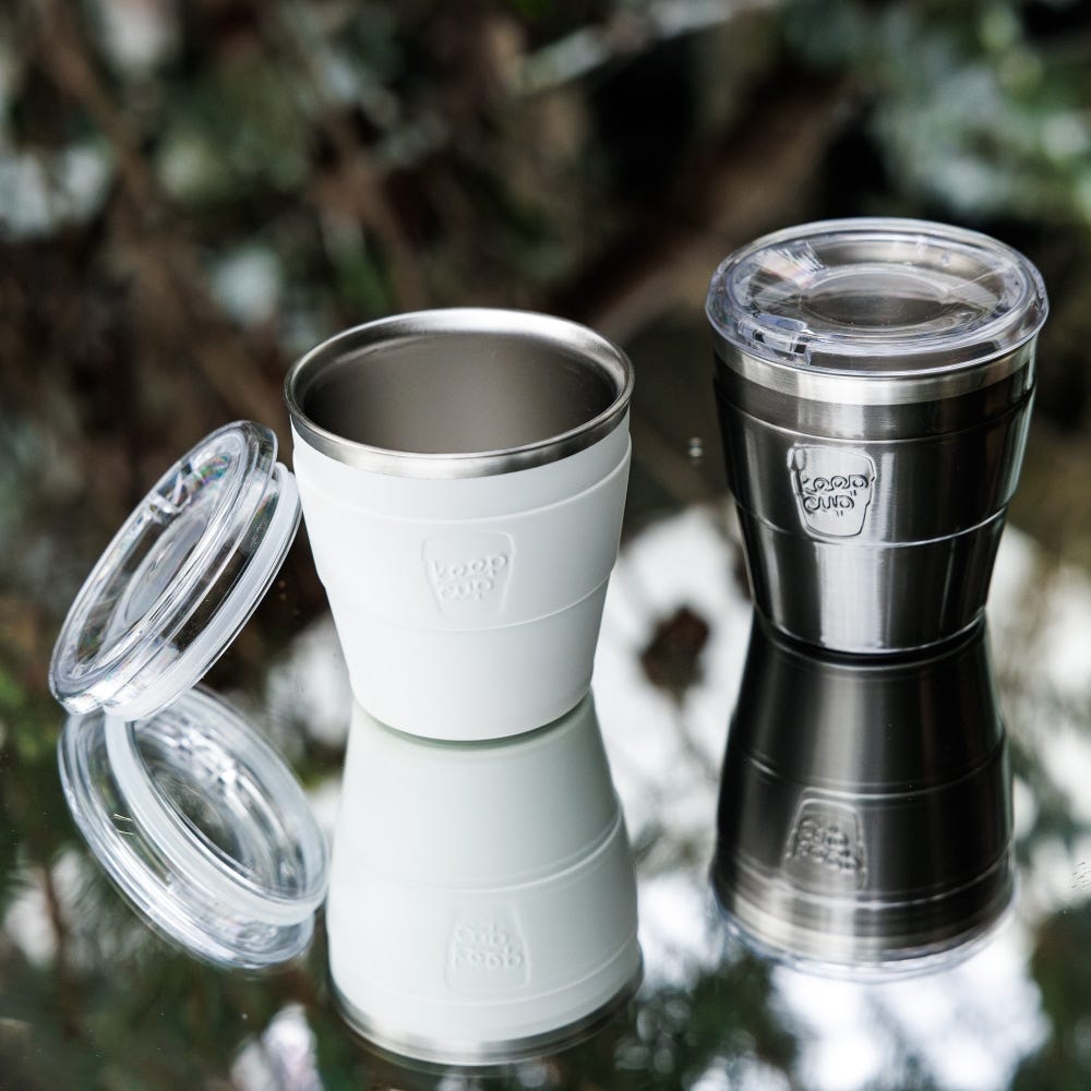 Two KeepCup reusable coffee cups with clear lids - one white, one black - displayed on reflective surface with blurred natural background