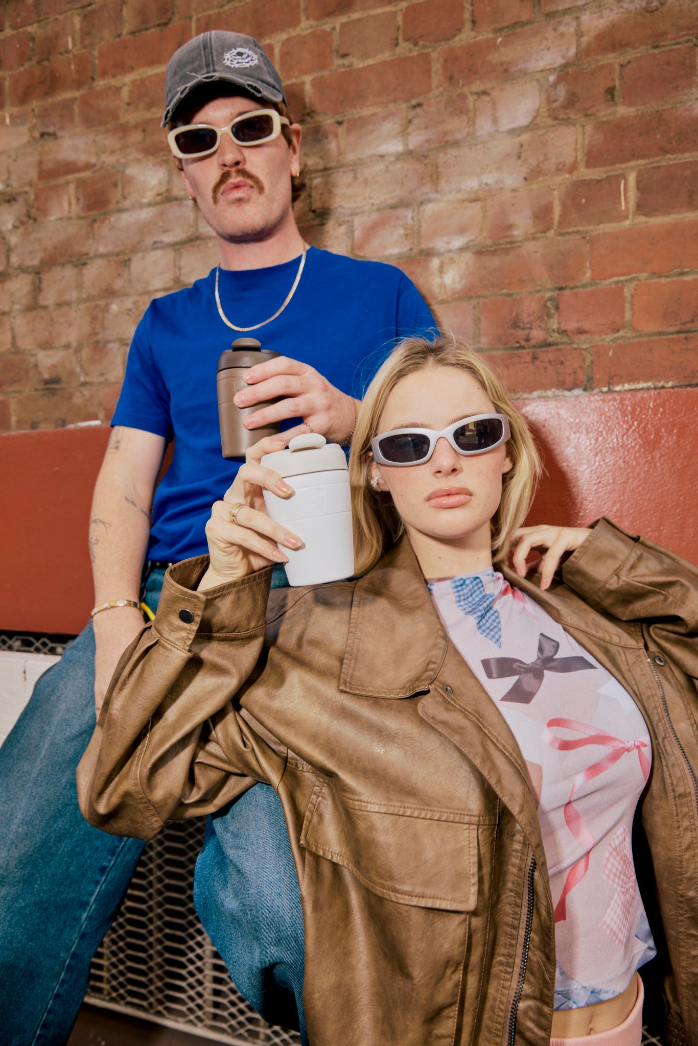 Two people sitting on a couch against a brick wall, holding drinks.