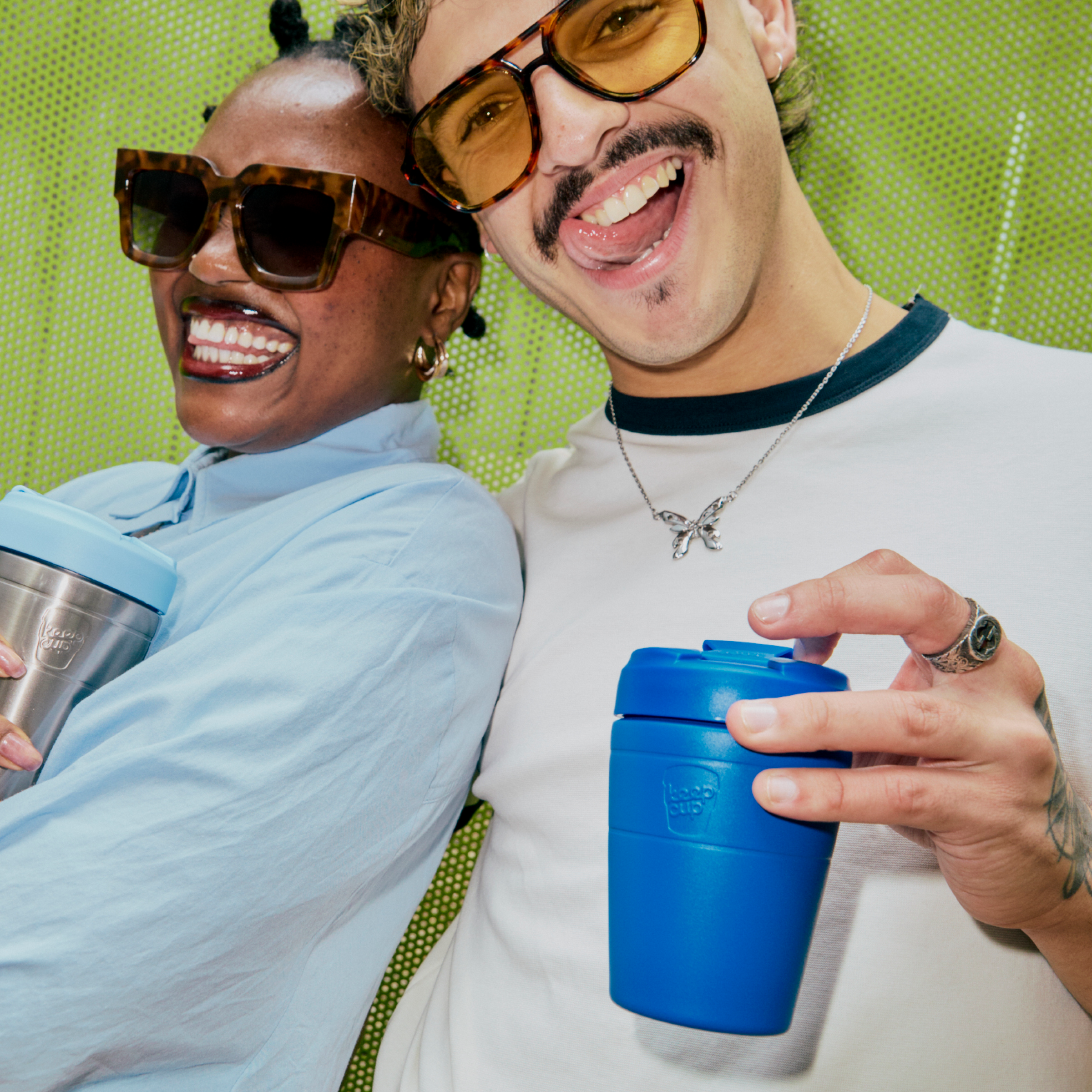 Two smiling people holding reusable KeepCup travel tumblers in a bright green perforated setting. One person wears tortoiseshell sunglasses and a light blue shirt, holding a silver tumbler with a light blue lid.