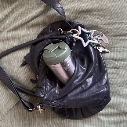 A black leather shoulder bag lying on a textured olive green fabric surface, with a silver insulated KeepCup travel mug and white keychain inserted into its side pocket.