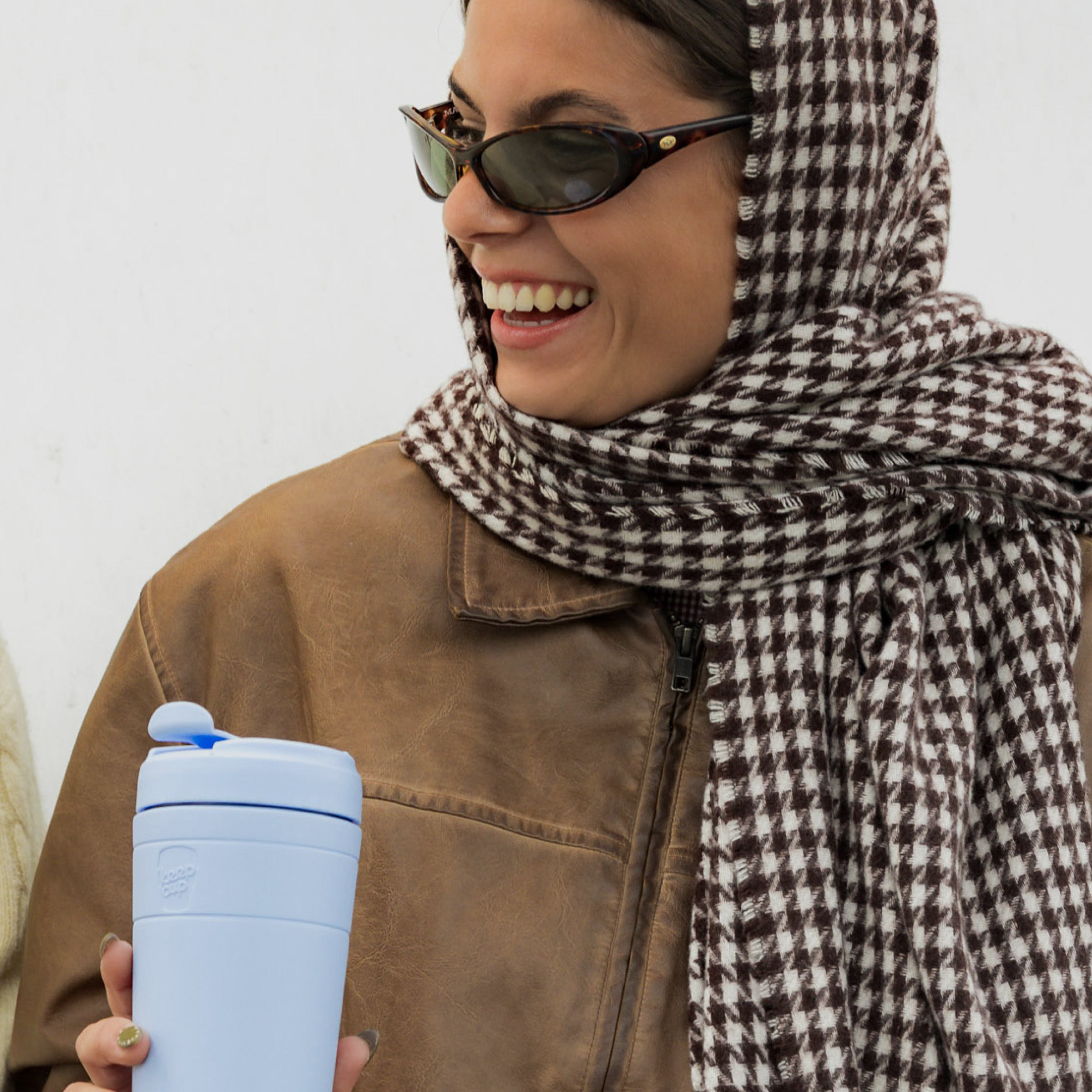A woman wearing a brown and white houndstooth patterned headscarf and sunglasses, smiling while holding a light blue KeepCup travel tumbler with a matching blue lid.