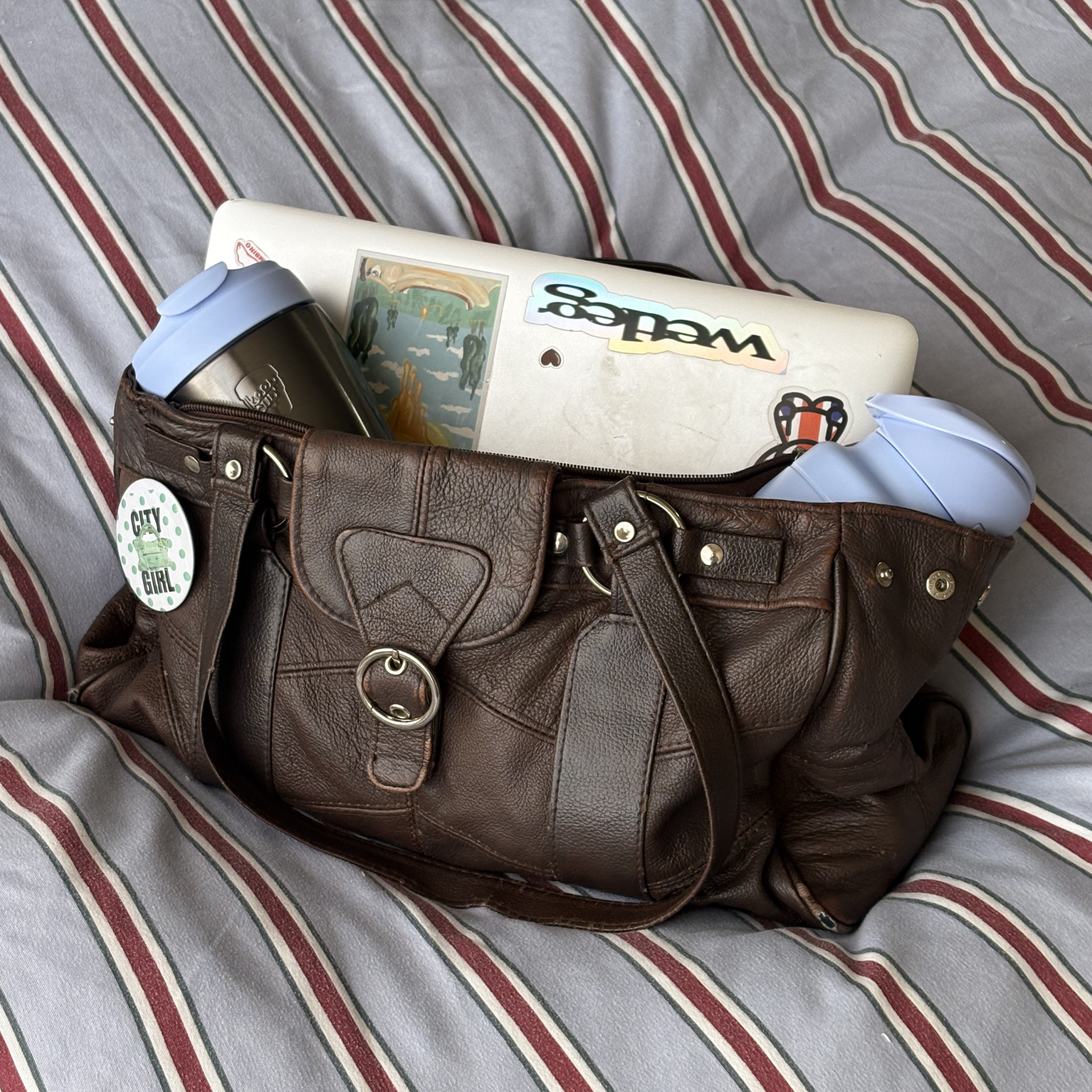 A brown leather shoulder bag filled with a laptop, two light blue KeepCup bottle and travel mug, resting on a gray and red striped bedsheet background.