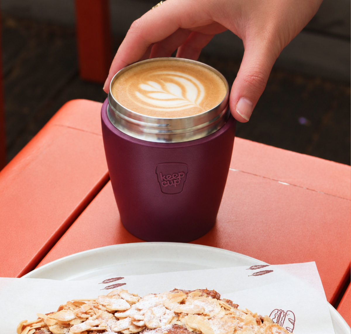A hand holds a Commuter Mulberry filled with latte coffee, on an orange table next to a plate of food.