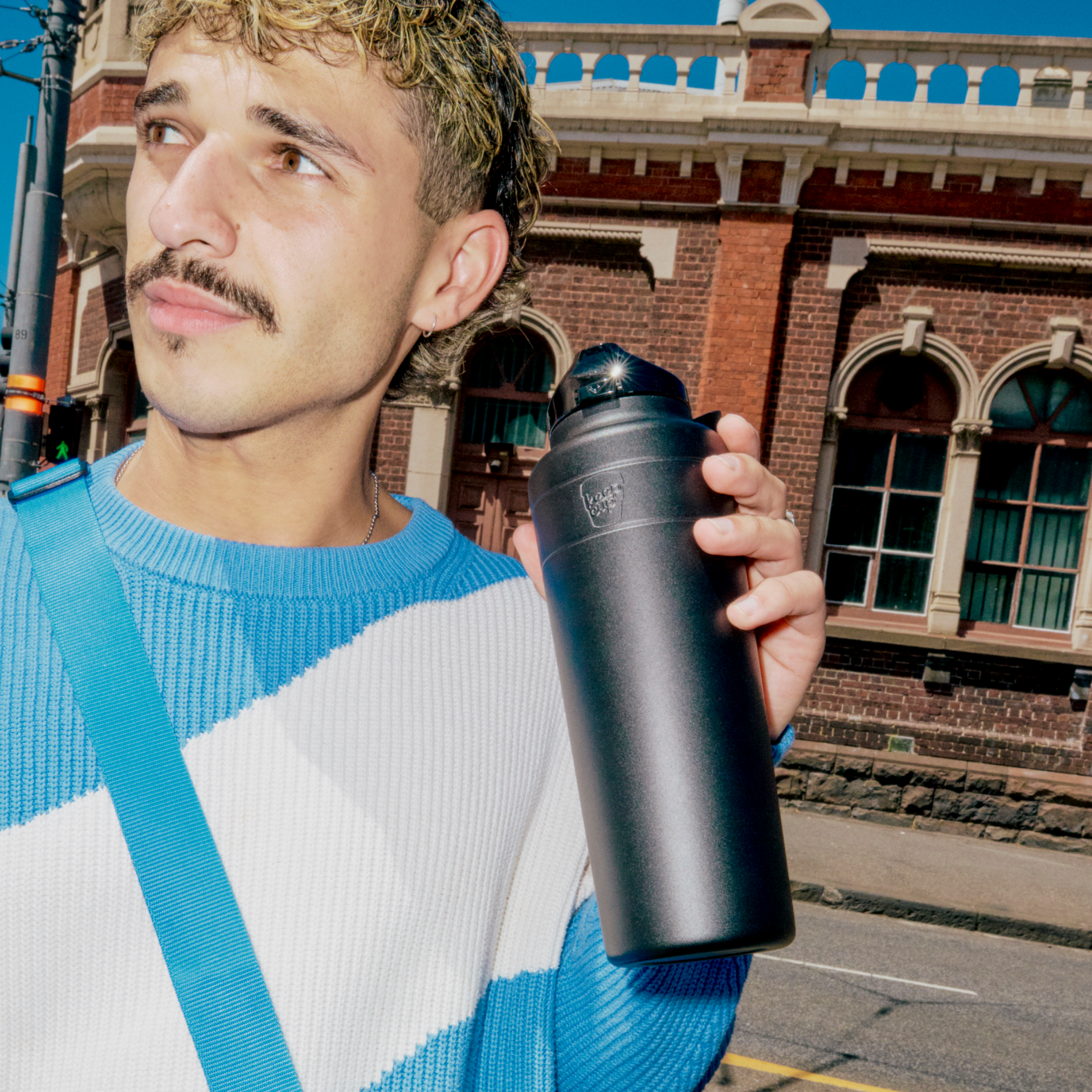 A man with curly hair and a moustache wearing a blue and white knit jumper holds a black Ora Bottle 950ml while standing on an urban street with a heritage brick building in the background.