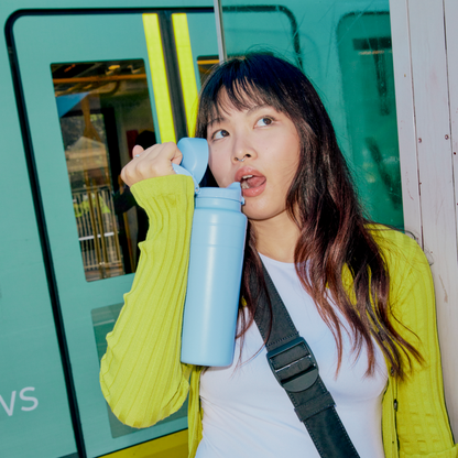 A young woman with long dark hair wearing a yellow cardigan drinks from a light blue KeepCup Ora Bottle while standing beside a teal train carriage, looking to the side mid-sip.