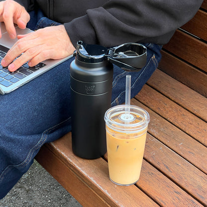 A person sits on a wooden bench working on a laptop, with a black KeepCup Ora Bottle 950ml beside them, its flip-top lid open and sunglasses resting on top, next to a clear reusable cup filled with iced coffee.