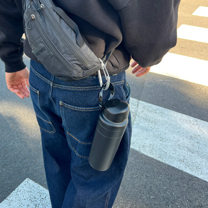 Person walking near a pedestrian crossing with a black Ora Bottle 950ml clipped to their belt loop via a carabiner, wearing dark denim jeans, a black hoodie and a grey Salomon crossbody bag.