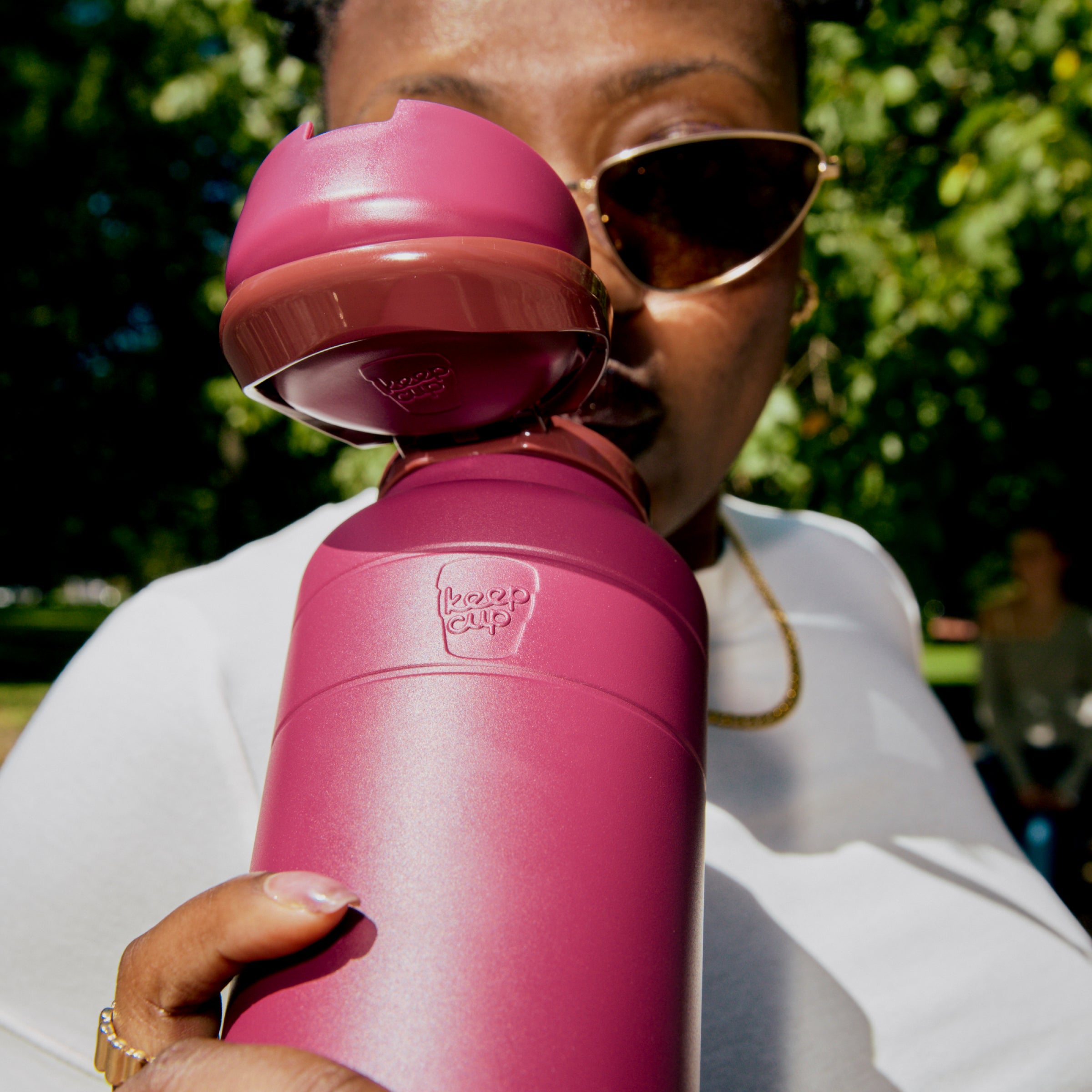 A woman wearing sunglasses and a white shirt holds up an Ora Bottle 950ml in Mulberry, with the flip-top lid open ready to drink, shown outdoors in a sunny park setting.