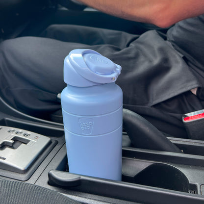 KeepCup Ora Bottle 700ml in Orchid, a matte periwinkle blue insulated bottle with flip-top lid, sitting in a car cup holder next to the gear shift.