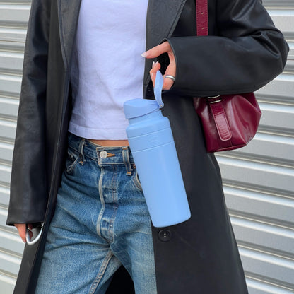 A woman in a black leather coat and blue jeans holds a KeepCup Ora Bottle 700ml in Orchid by its carry loop, showcasing the light blue reusable bottle against a corrugated metal backdrop.