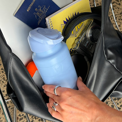 A hand with silver rings places an Ora Bottle 700ml in Orchid into a black leather tote bag alongside travel essentials including an Australian passport, yellow notebook, Panasonic Lumix camera, and orange sunscreen bottle.