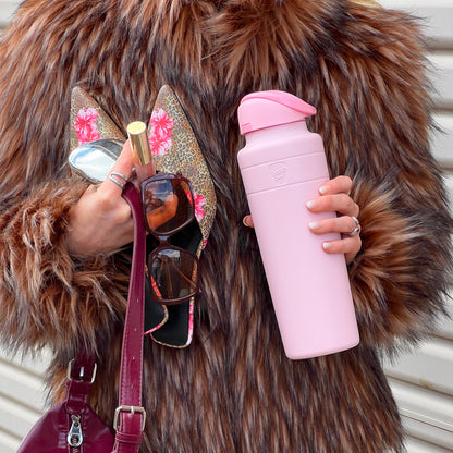 A person wearing a fluffy brown faux fur coat holds the KeepCup Ora Bottle 700ml in Pashmak Pink in one hand, while the other hand holds leopard print heels with pink floral details, sunglasses, and a gold lipstick, with a burgundy handbag strap visible.