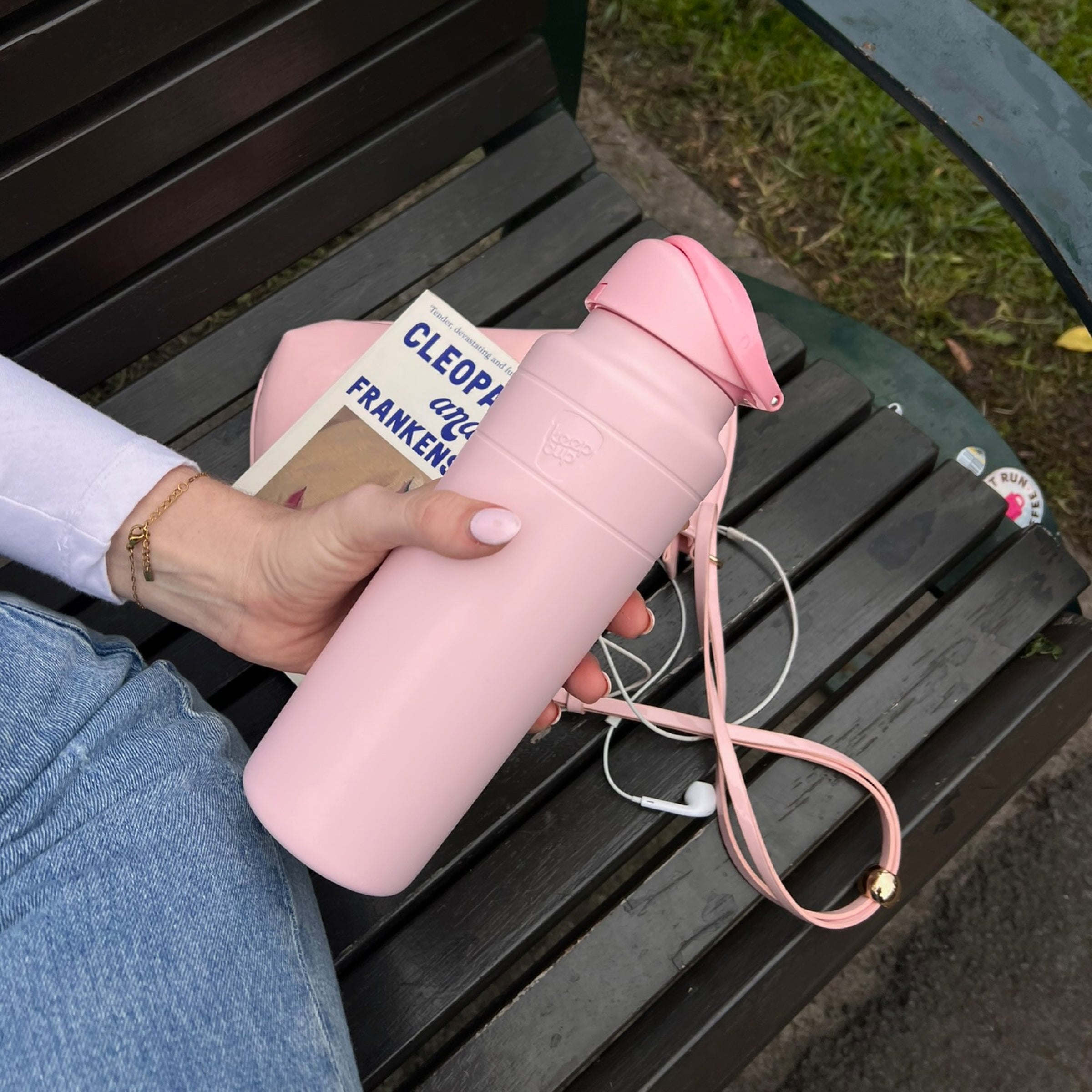A hand with white nail polish holds the KeepCup Ora Bottle 700ml in Pashmak Pink while sitting on a dark wooden park bench, with a paperback book, white earphones and the bottle's pink carry strap visible alongside.